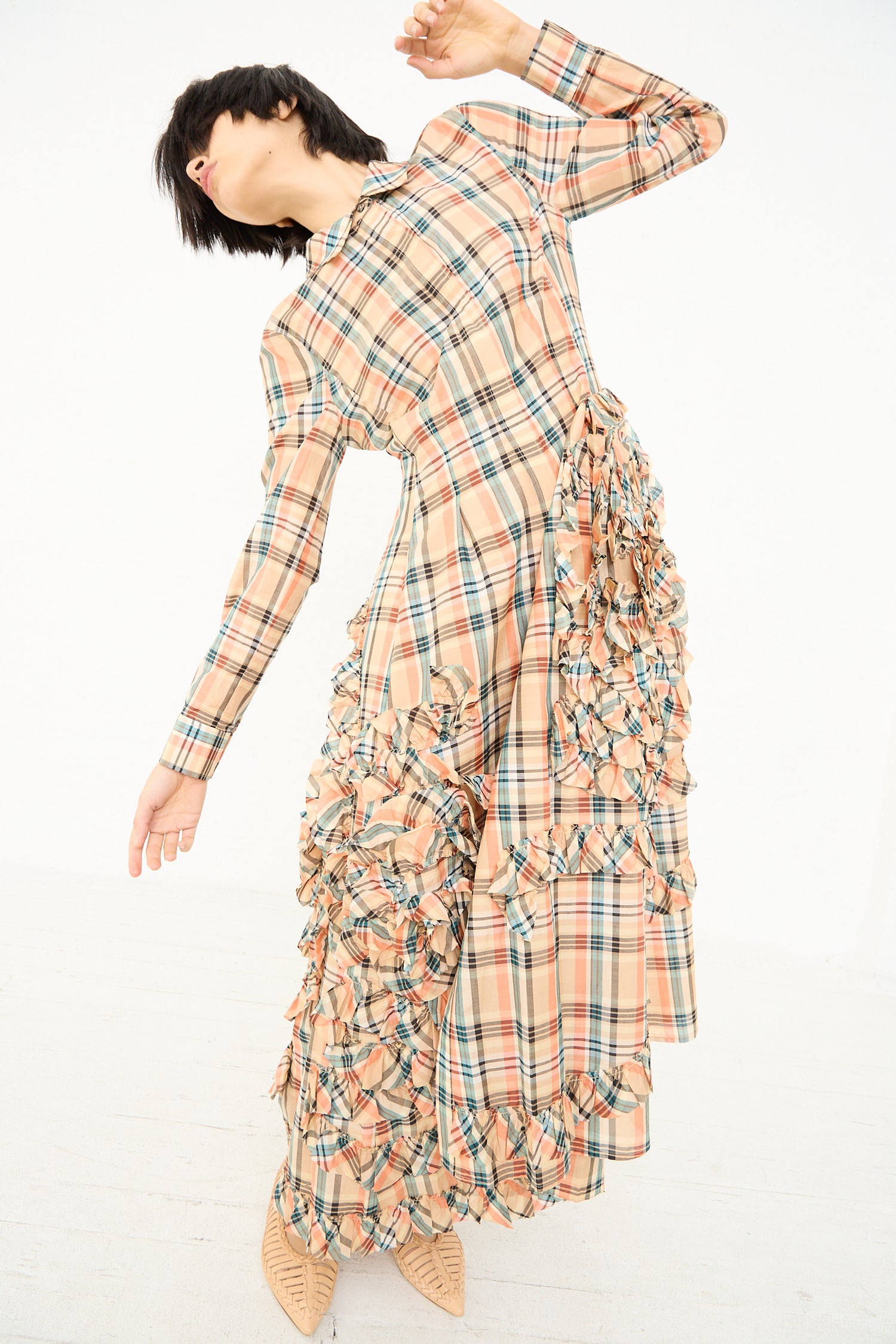 A person in a Ulla Johnson Gwen Dress in Meadow, a plaid maxi shirt dress with ruffle details, mid-motion with head tilted back and arm extended.