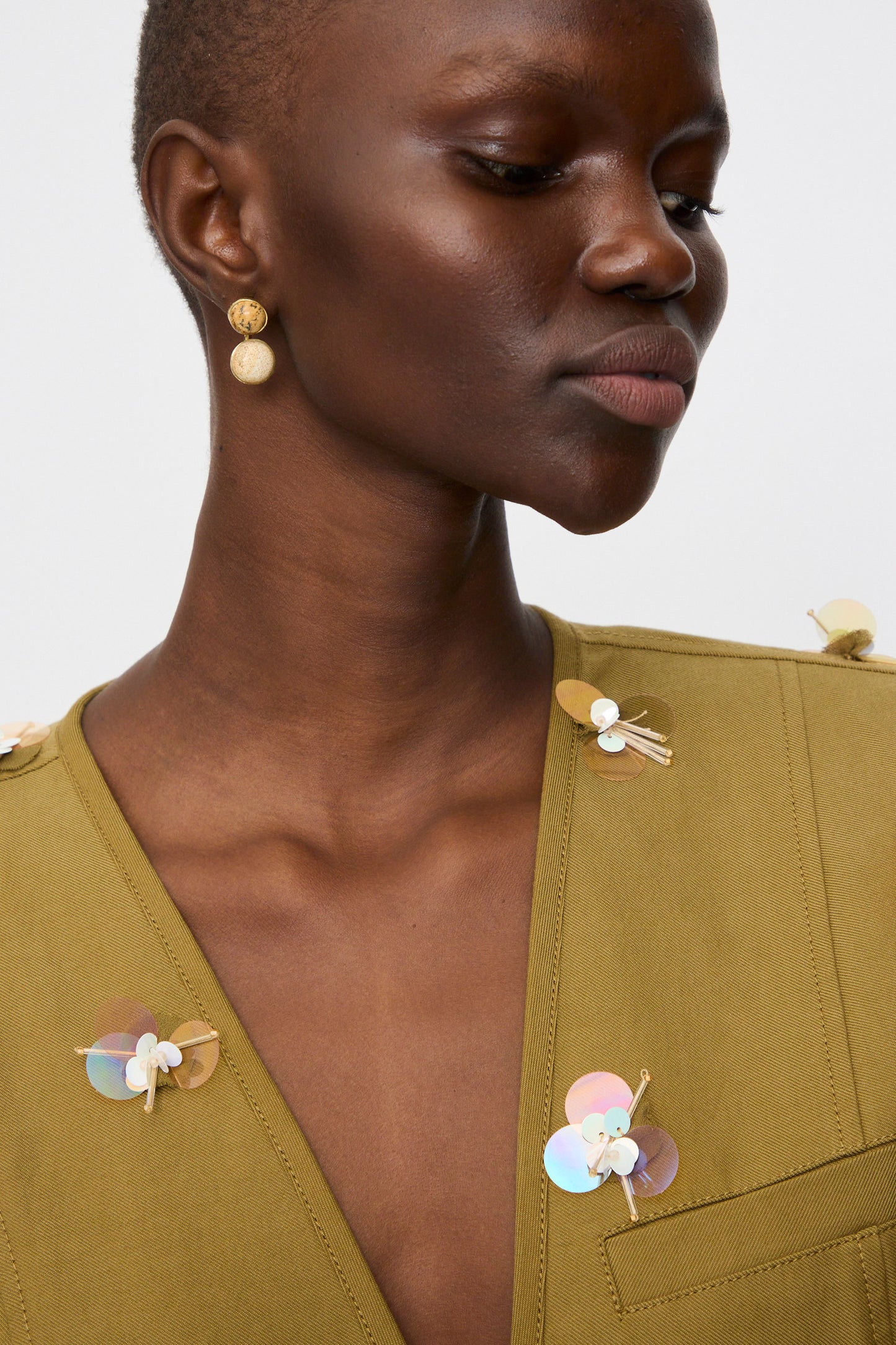 Person wearing a light olive green top with large iridescent sequin and bead embellishments, accessorized with Ulla Johnson's Kiri Double Stone Earring in Picture Jasper, and looking downward.