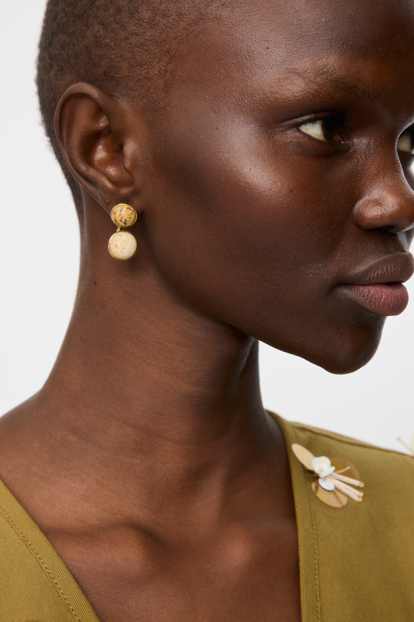 Close-up of a person wearing Ulla Johnson's Kiri Double Stone Earring in Picture Jasper, paired with an olive green top featuring a decorative shoulder pin.