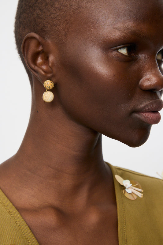 Close-up of a person wearing Ulla Johnson's Kiri Double Stone Earring in Picture Jasper, paired with an olive green top featuring a decorative shoulder pin.