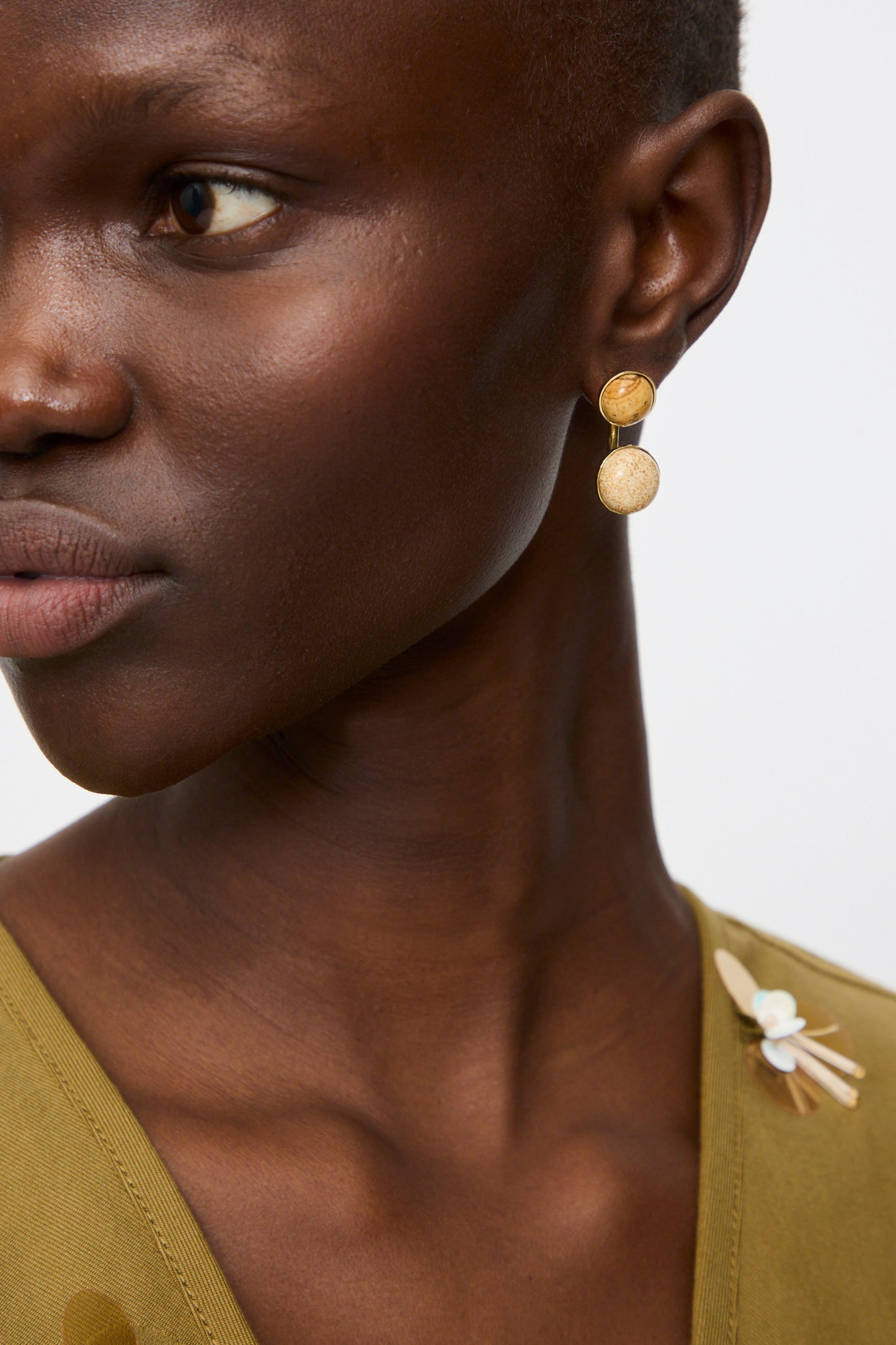Close-up of a person wearing Ulla Johnson’s Kiri Double Stone Earring in Picture Jasper and an olive green top with a decorative shoulder pin, set against a plain background.