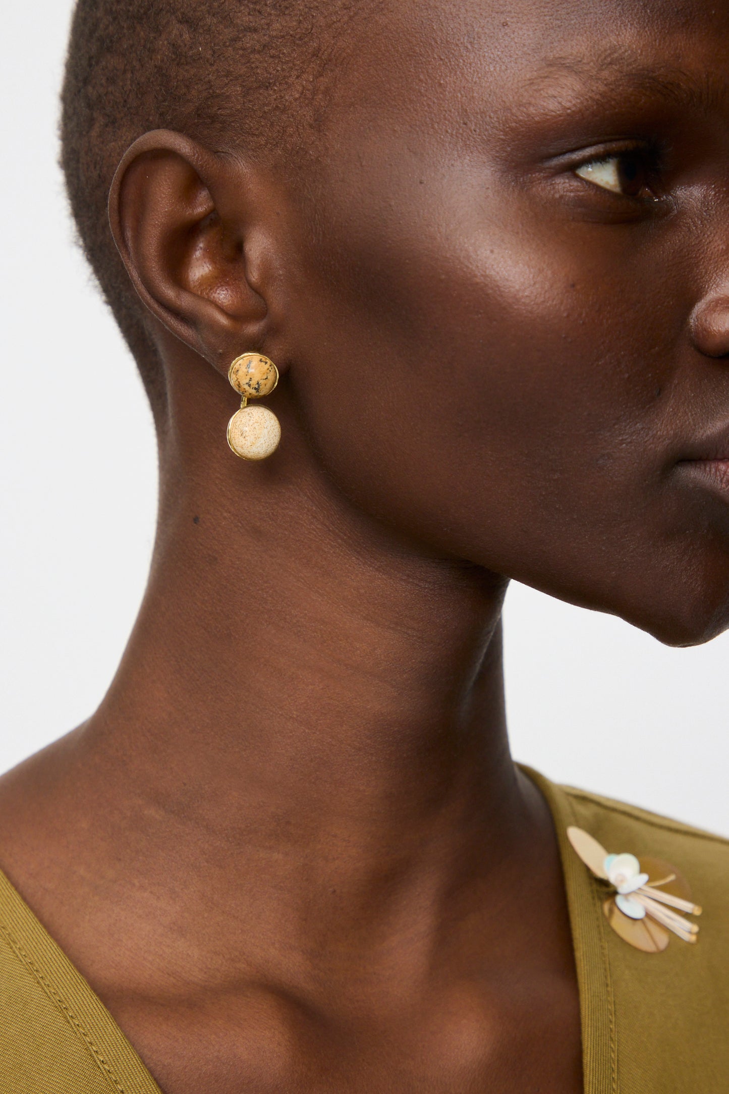 A person wearing a green top styled with Ulla Johnson's Kiri Double Stone Earrings in Picture Jasper and a pin decorated with a small blue flower and beige leaves.