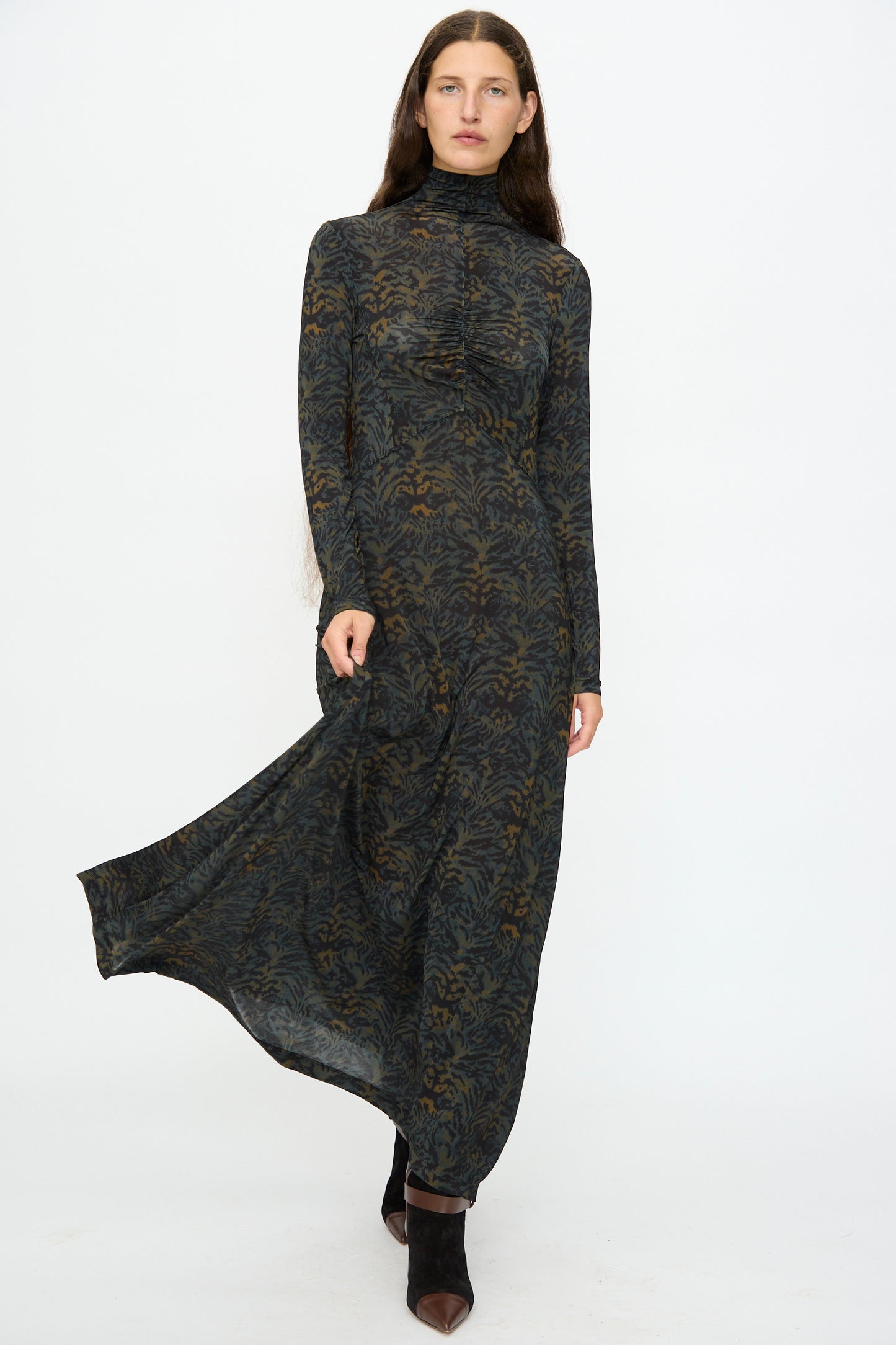 A woman stands against a plain background wearing the Ulla Johnson Koa Dress in Desert Jasper, a slim fit maxi with long sleeves and high neck, featuring a dark ikat animal print. She pairs it with black pointed-toe shoes and lifts the hem slightly.