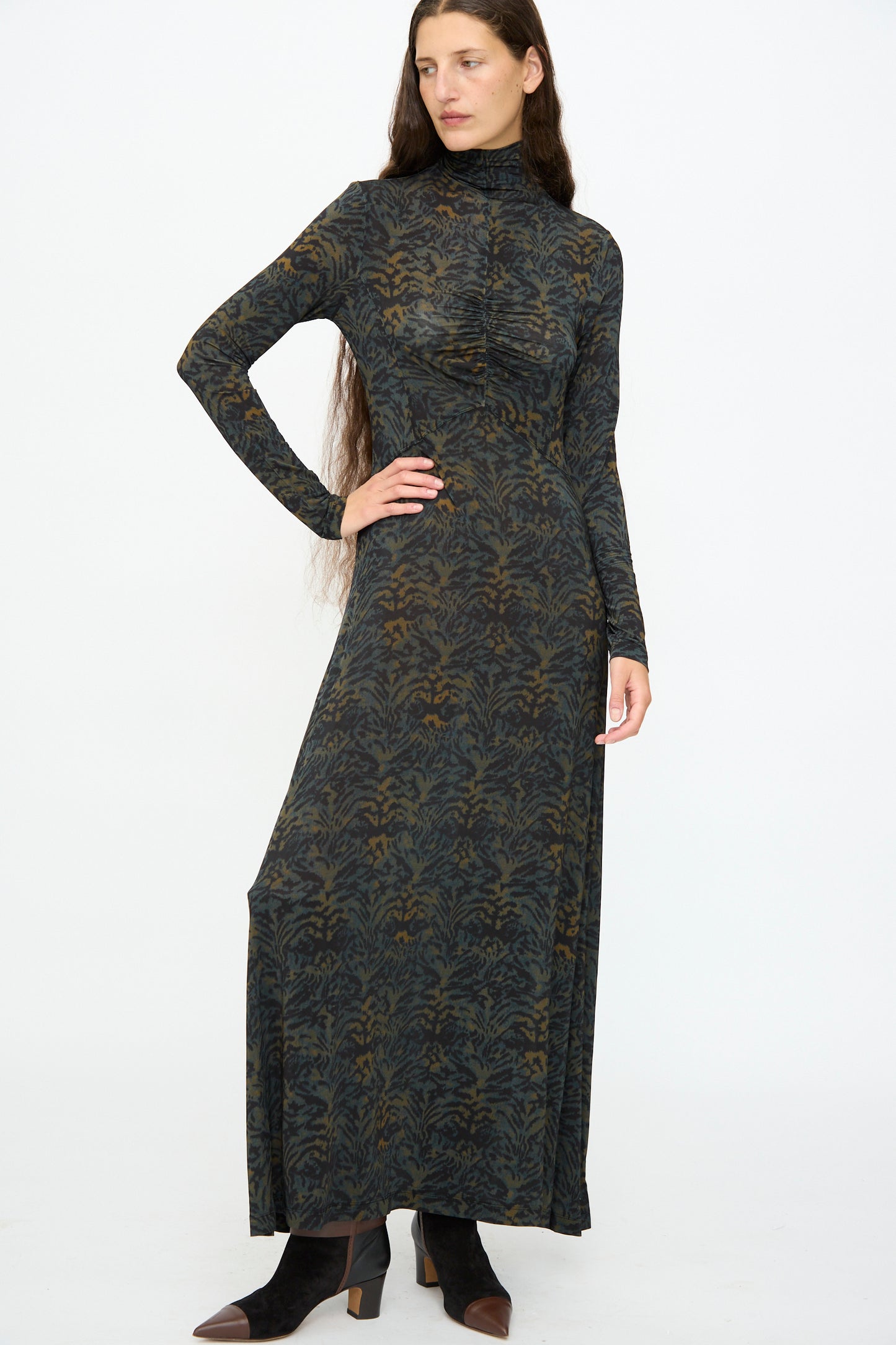 A woman wears the Ulla Johnson Koa Dress in Desert Jasper, a slim fit maxi with ikat animal print, long sleeves, and a high neckline, paired with black ankle boots against a plain white background.