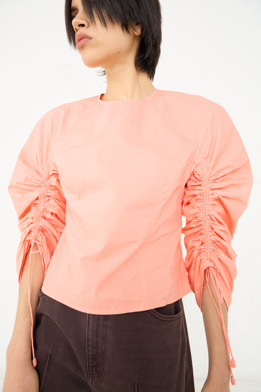 Woman in a Lorna Blouse in Peony by Ulla Johnson, a coral pink cotton poplin blouse with ruched sleeves, standing against a white background.