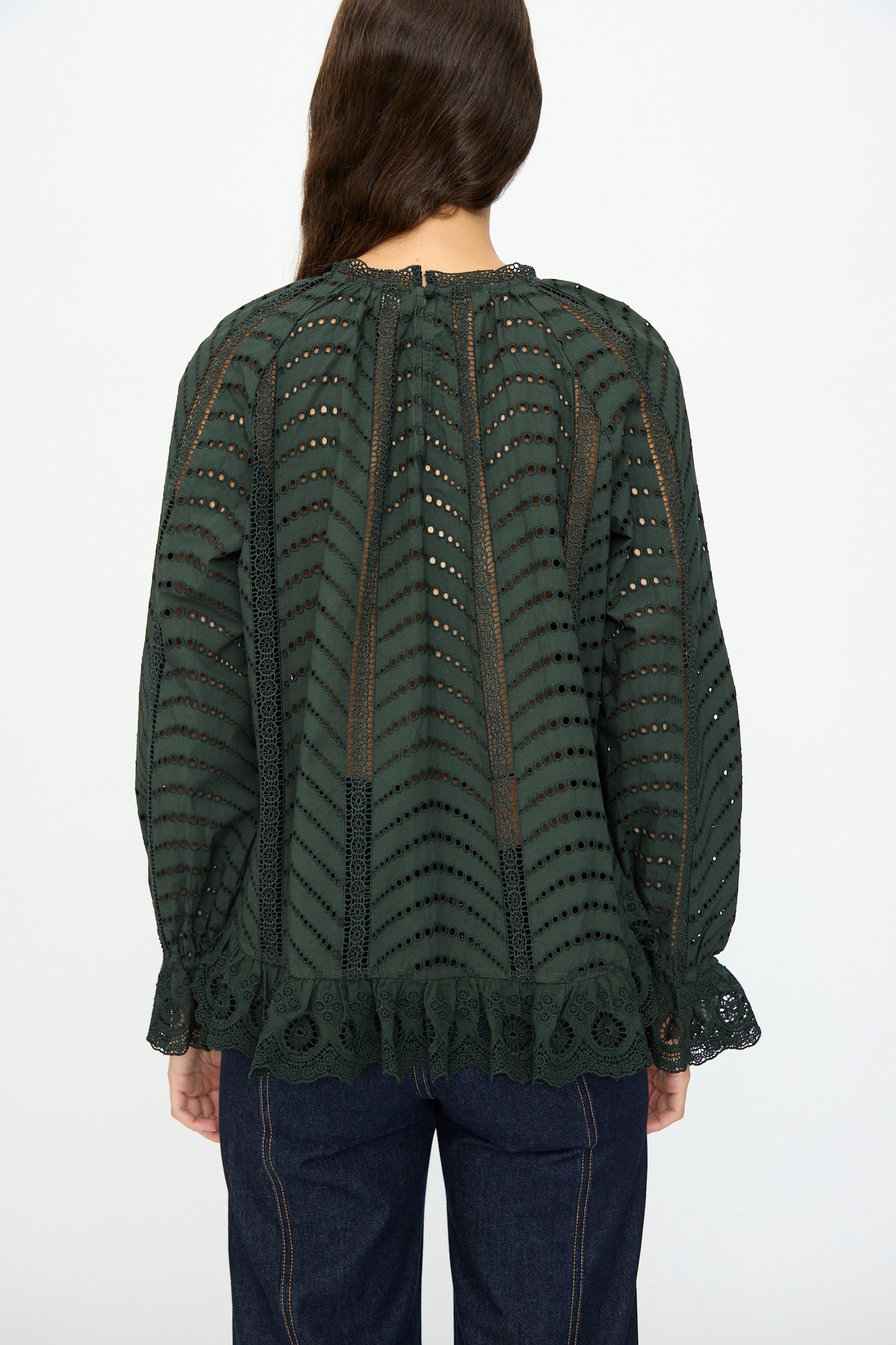 A person with long brown hair is photographed from the back, wearing the Ulla Johnson Marcella Blouse in Cypress—a dark green, long sleeve cotton top with eyelet details and scalloped ruffles—paired with dark blue jeans.