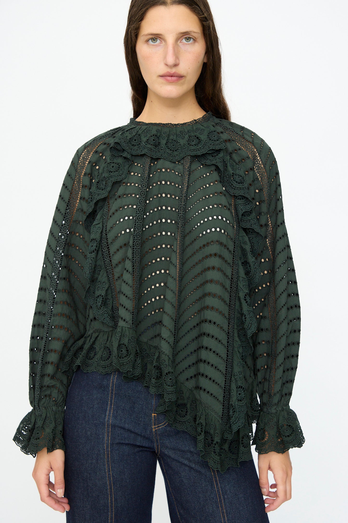 A woman wearing the Ulla Johnson Marcella Blouse in Cypress, featuring long sleeves, lace, and scalloped ruffles, paired with dark blue jeans against a plain white background.