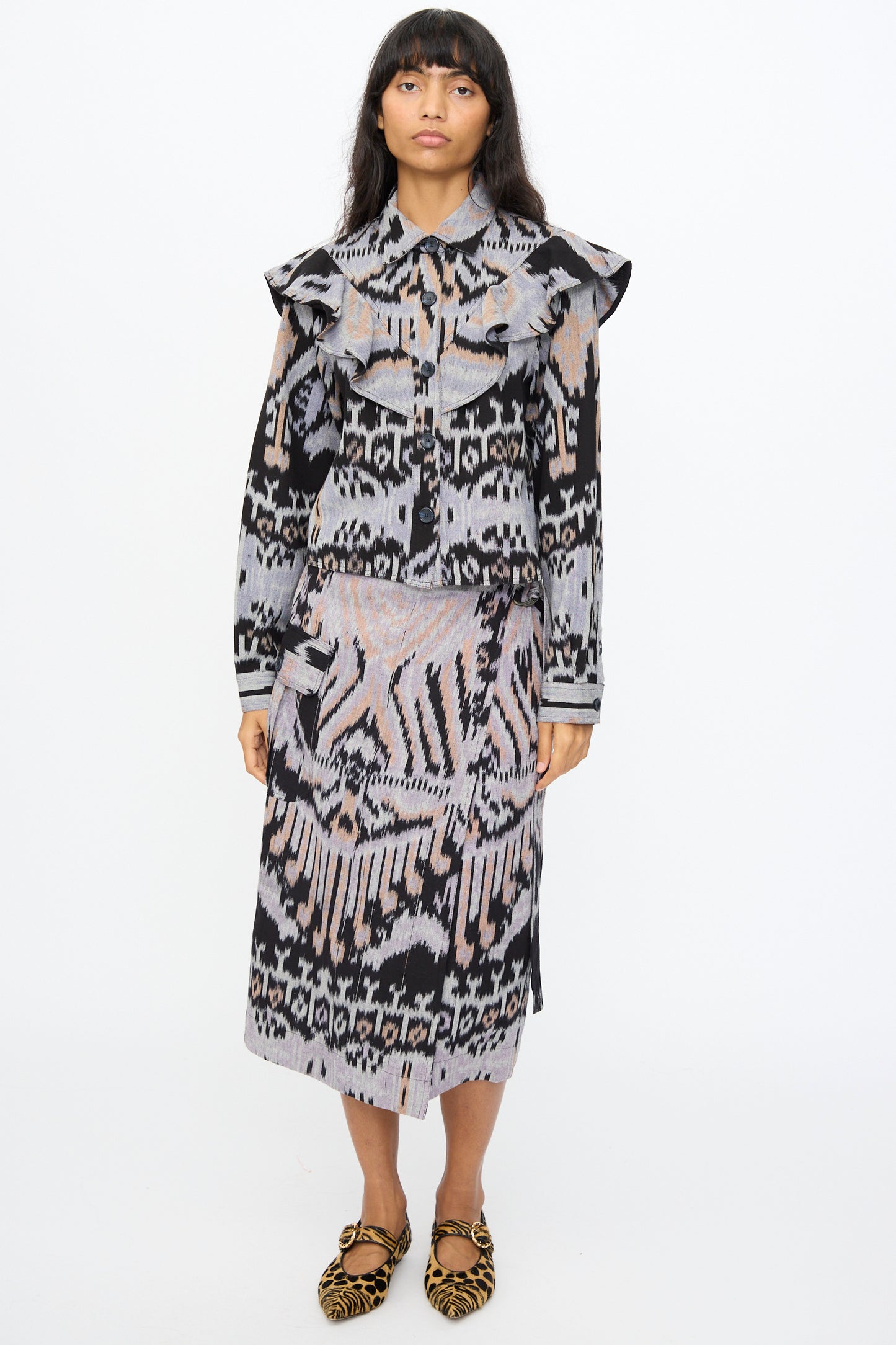A person stands facing forward in the Ulla Johnson Mathilde Blouse in Tigereye, featuring hand-loomed cotton with ikat geometric designs in black, gray, and beige, paired with a matching skirt and brown animal print shoes against a plain white background.