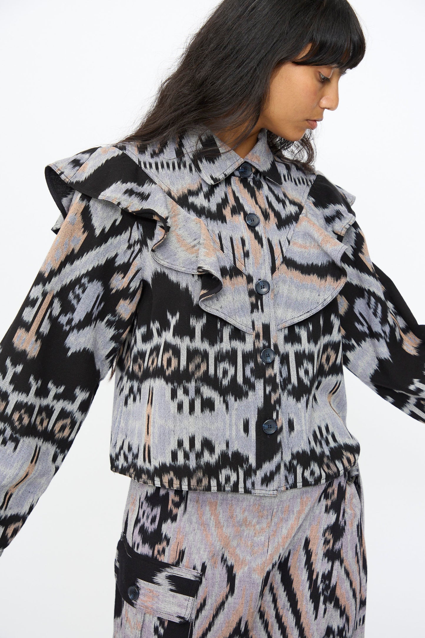 A woman wears the Ulla Johnson Mathilde Blouse in Tigereye, a patterned button-up with ruffled shoulders featuring abstract black, gray, and peach Ikat motifs for a unique look. Crafted in India.