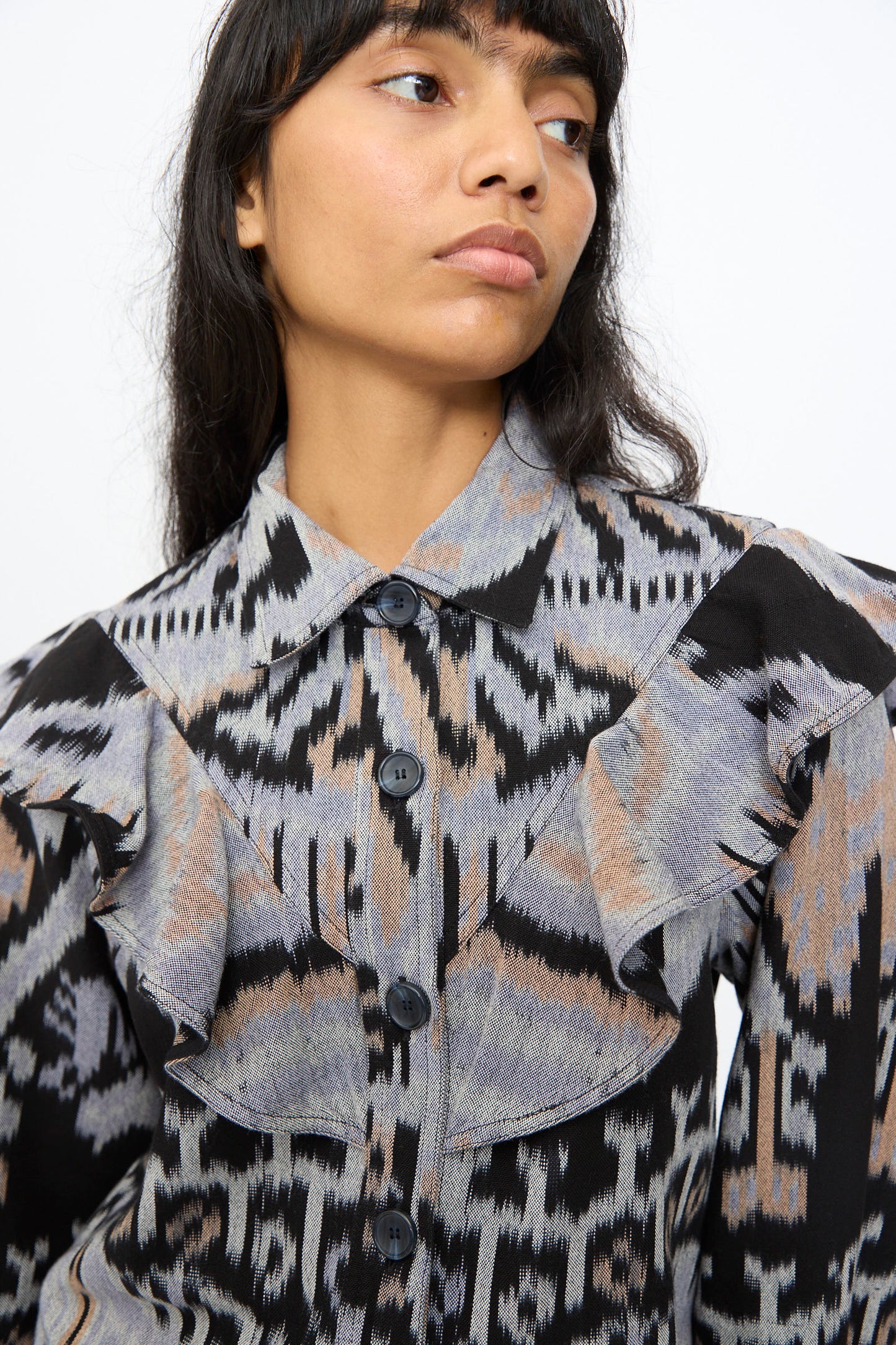 Someone wearing the Ulla Johnson Mathilde Blouse in Tigereye—a hand-loomed cotton top featuring a bold geometric Ikat pattern and ruffled front—looks slightly to the side.