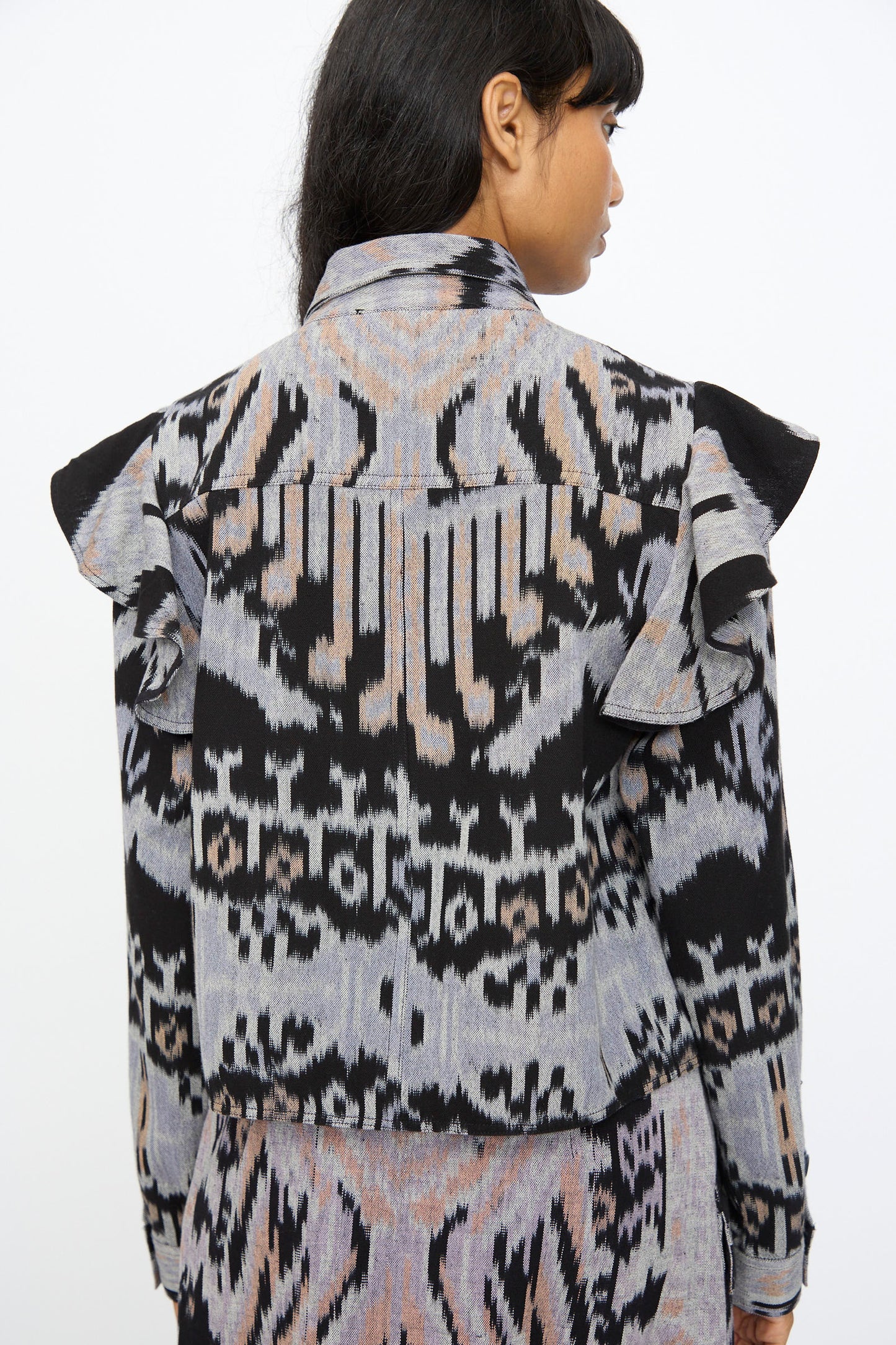 A woman is seen from behind wearing the Ulla Johnson Mathilde Blouse in Tigereye, a hand-loomed cotton top with long sleeves, ruffled shoulders, and a geometric black, gray, and beige Ikat design.