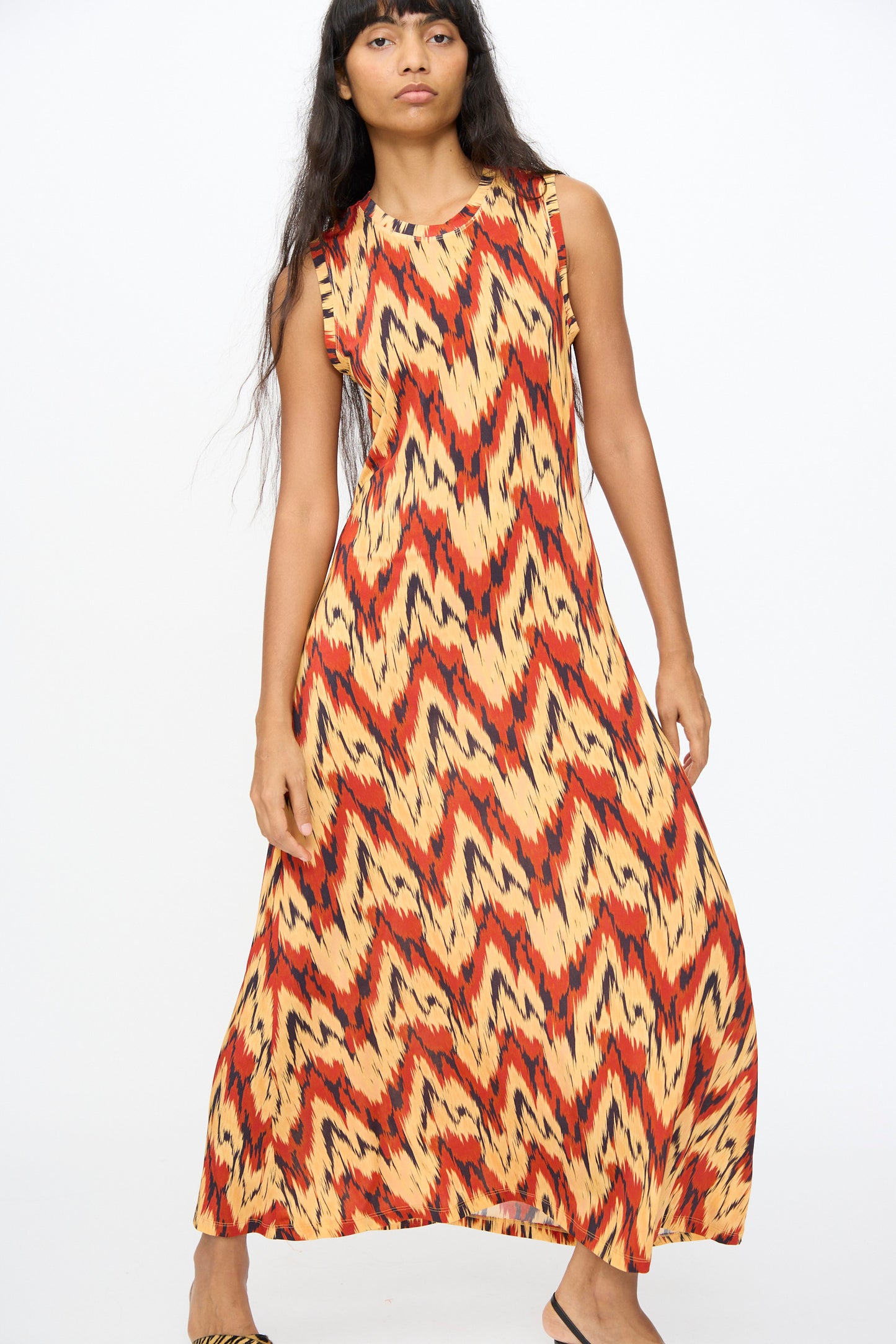 A woman models the Ulla Johnson Orla Dress in Carnelian, a sleeveless tricot jersey dress with a bold zigzag pattern in yellow, orange, and black, against a plain white background.