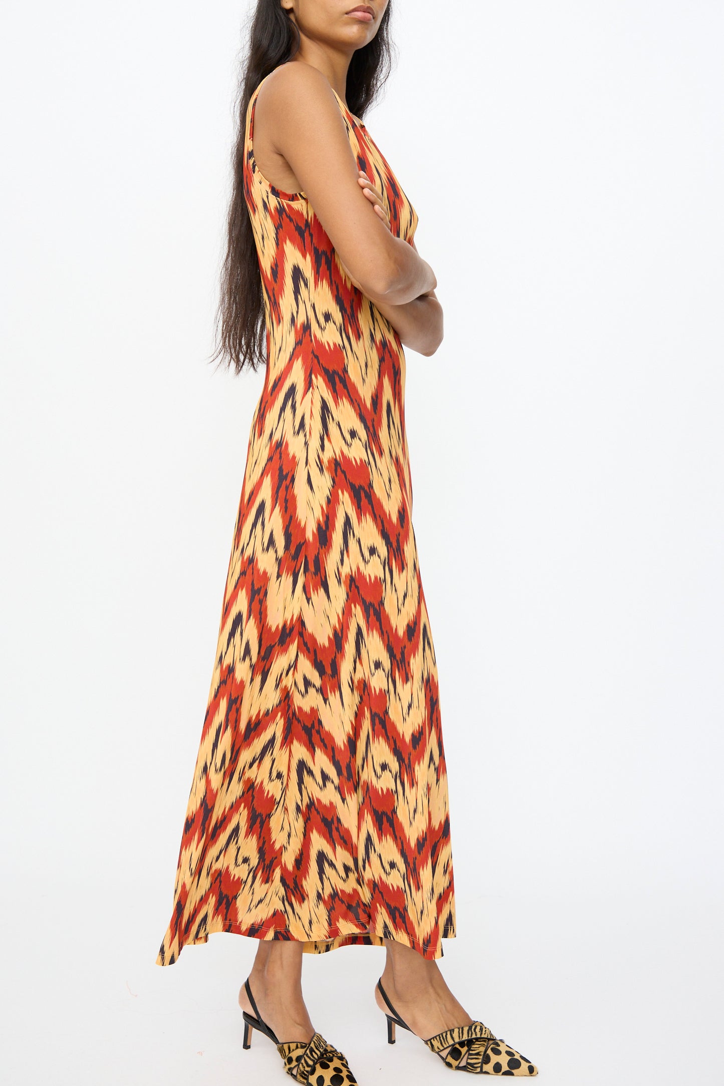 A woman stands in profile with arms crossed, wearing the Ulla Johnson Orla Dress in Carnelian—crafted from tricot jersey with a red, yellow, and black abstract pattern—and striking leopard print heels.