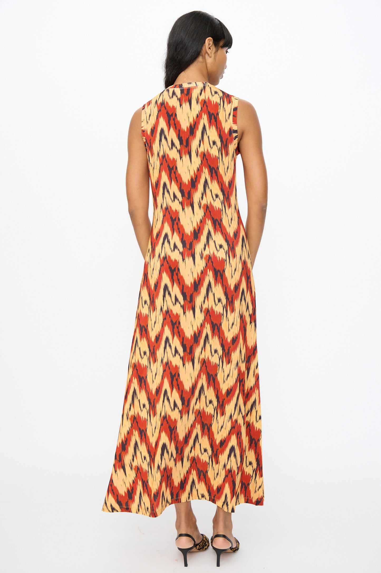 A person stands with their back to the camera, wearing the Ulla Johnson Orla Dress in Carnelian—a sleeveless, ankle-length tricot jersey dress with a bold red, yellow, and black zigzag pattern.