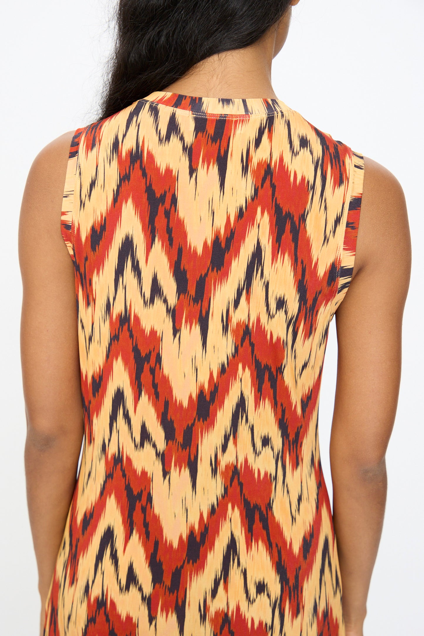 A person shown from the back wears the Ulla Johnson Orla Dress in Carnelian, a sleeveless dress featuring a red, yellow, and black zigzag tricot jersey pattern.