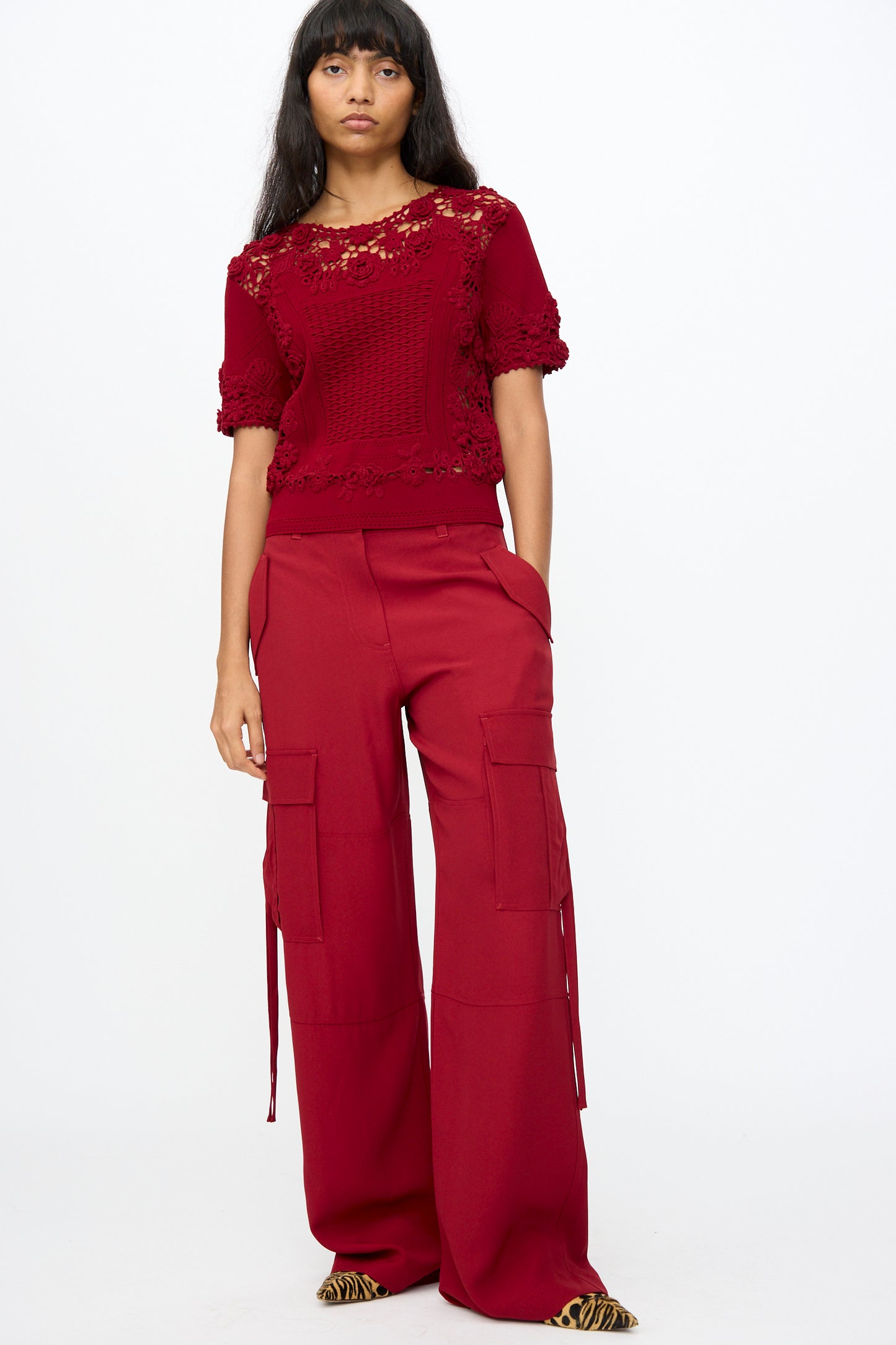 A person stands wearing the Pax Pant in Dahlia by Ulla Johnson—a relaxed fit, wide-leg cargo pant—paired with a red short-sleeve lace top and pointed tiger stripe shoes.