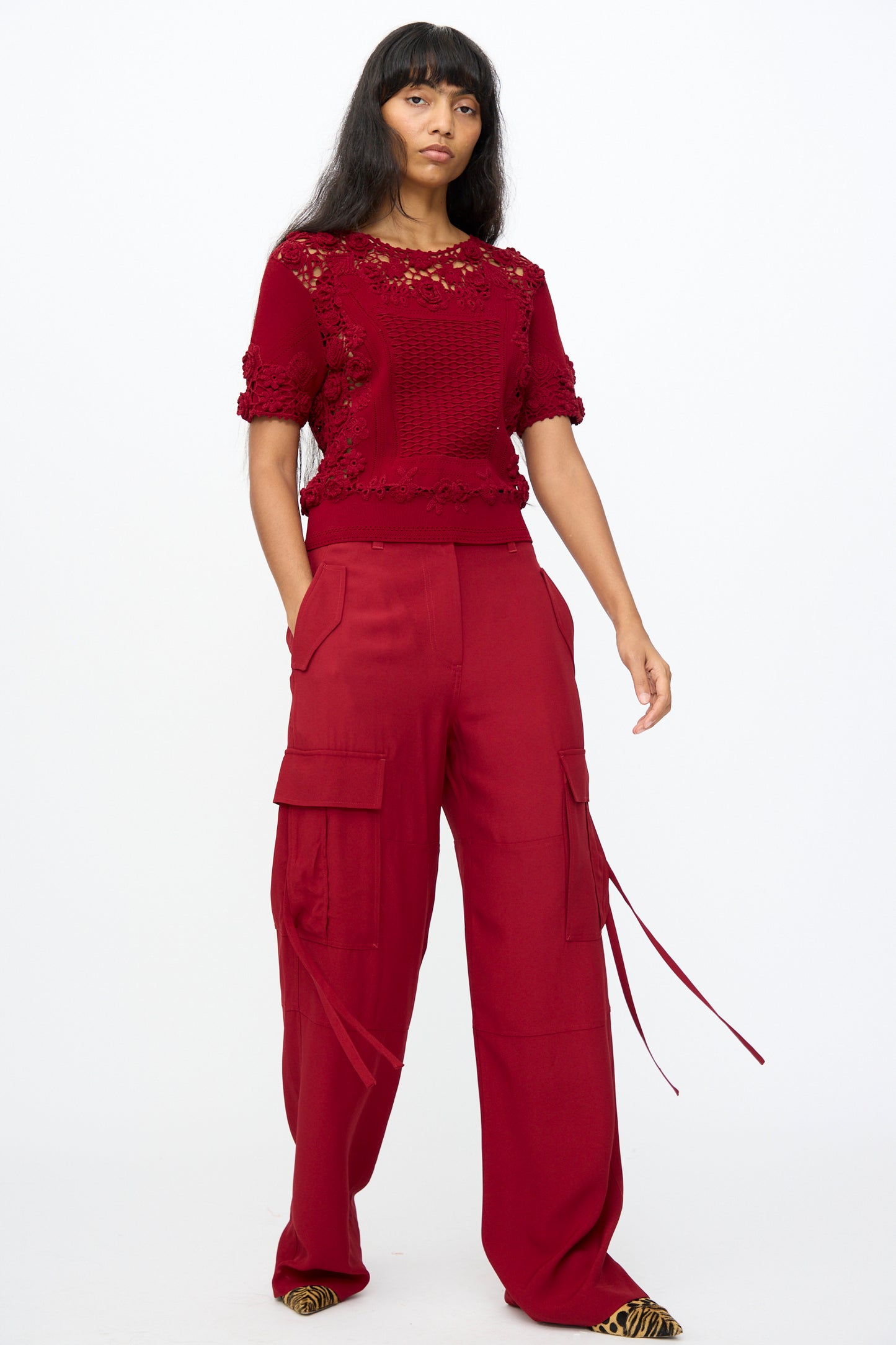 A woman poses in Ulla Johnson's Pax Pant in Dahlia, styled with a red lace short-sleeve top and animal print pointed shoes, against a plain white background.