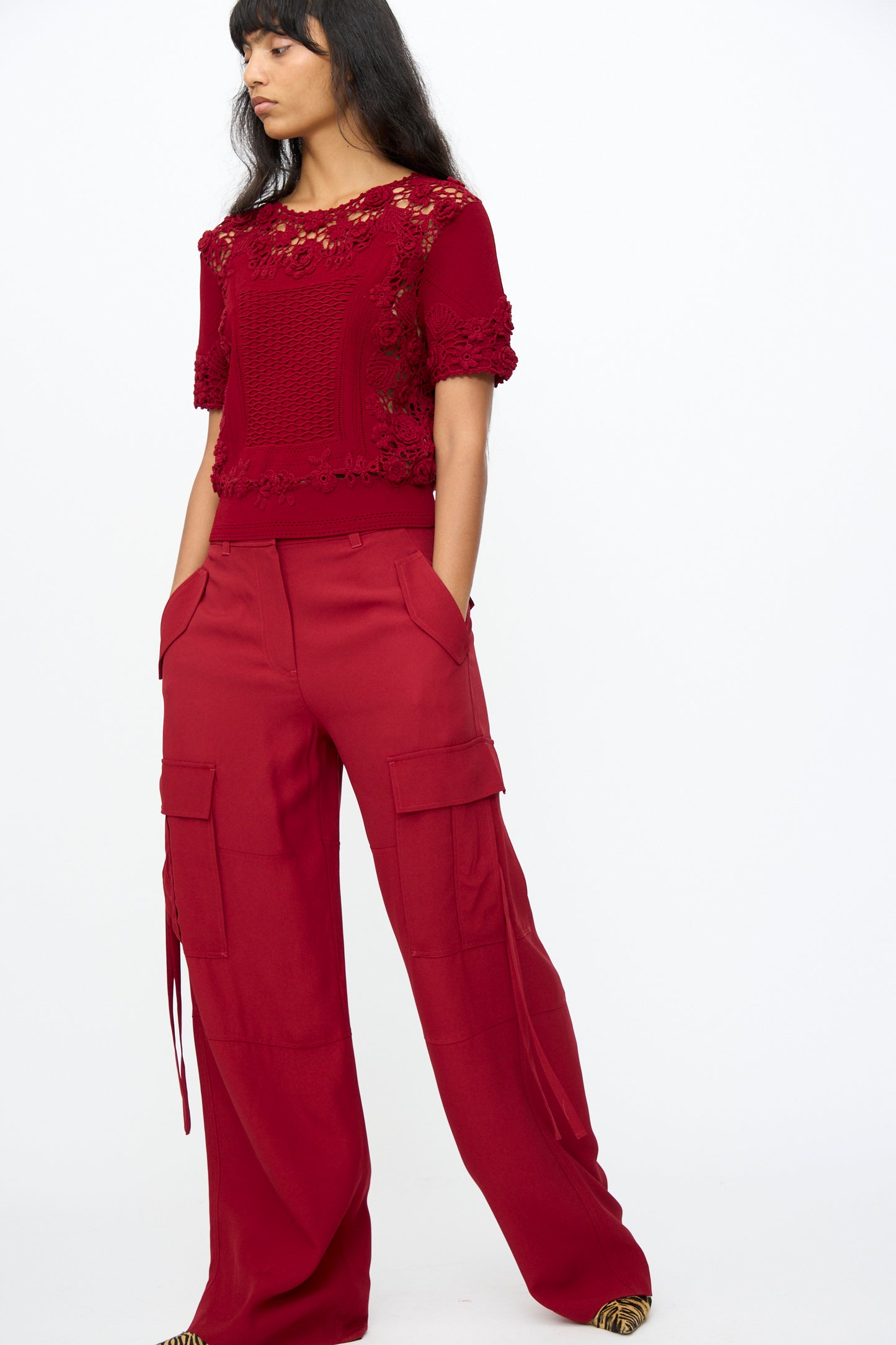 Against a plain background, a woman wears the Ulla Johnson Pax Pant in Dahlia—relaxed fit red cargo pants—with her hands in her pockets and pairs them with a red lace short-sleeve top.