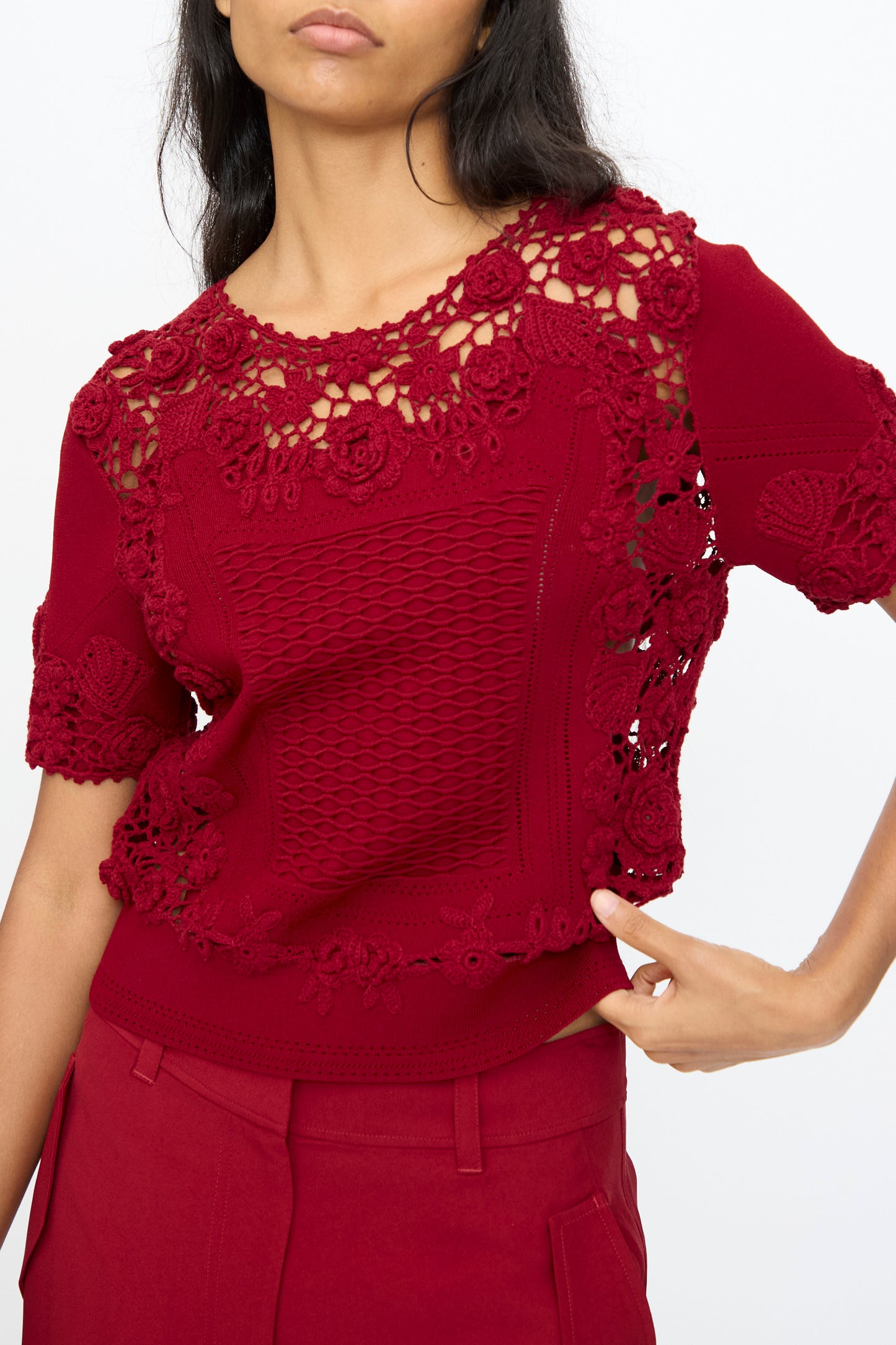 A woman stands against a plain light background wearing the Ulla Johnson Stella Knit Top in Begonia, a short-sleeved, dark red floral knit with a scalloped hem, paired with matching red pants.