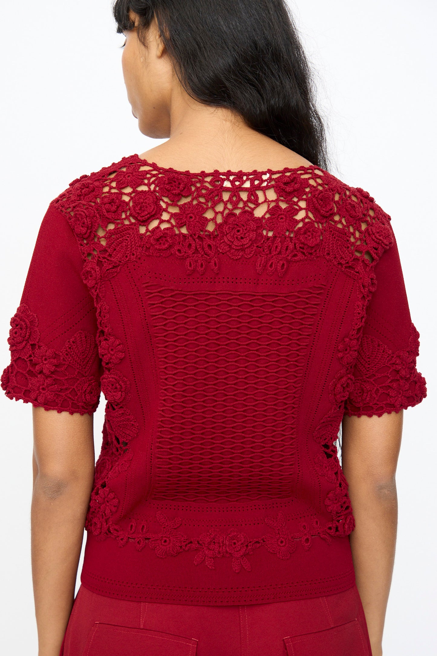 A woman is seen from the back wearing the Ulla Johnson Stella Knit Top in Begonia, a short sleeve style with intricate crochet and lace details on the shoulders, sleeves, and upper back, finished with a scalloped hem.