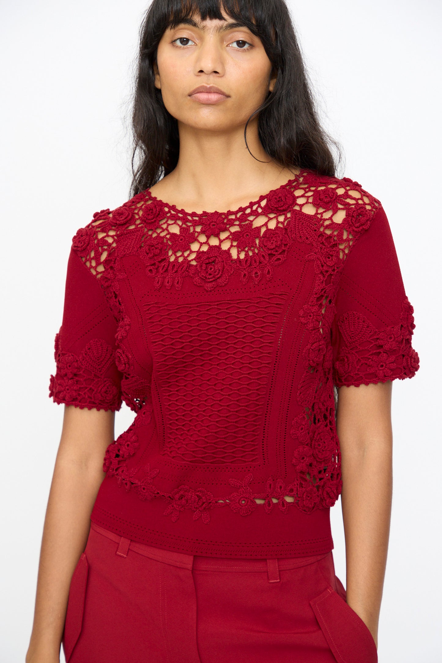 A woman stands against a plain light background wearing the Ulla Johnson Stella Knit Top in Begonia, a red short sleeve crochet lace top with a scalloped hem, paired with matching red pants.