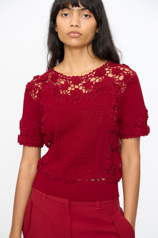 A woman stands against a plain light background wearing the Ulla Johnson Stella Knit Top in Begonia, a red short sleeve crochet lace top with a scalloped hem, paired with matching red pants.