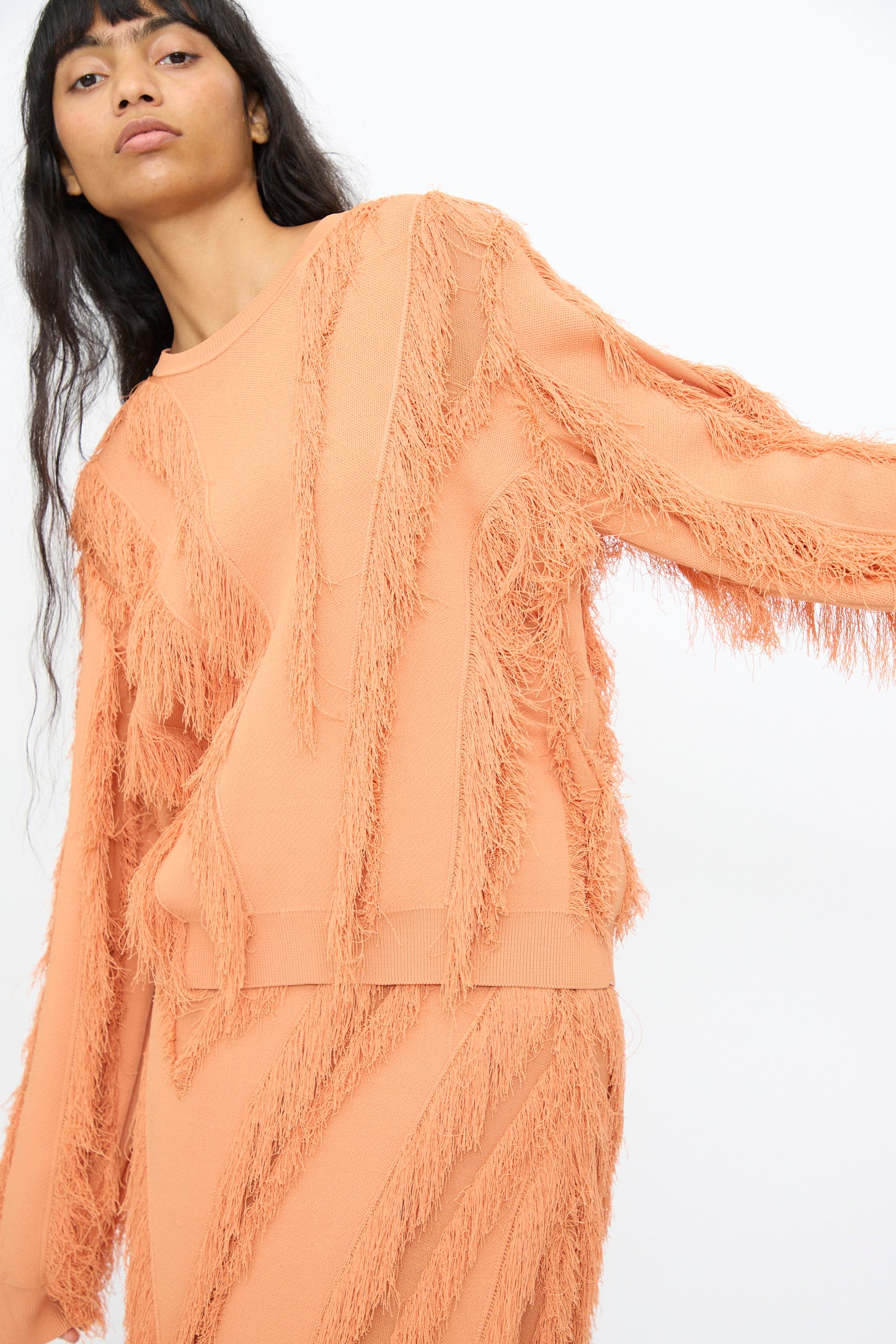 A person wearing the Ulla Johnson Aubree Knit Pullover in Tulip, a peach-colored, long-sleeve sweater with a fringe pattern and matching skirt, stands against a plain white background.