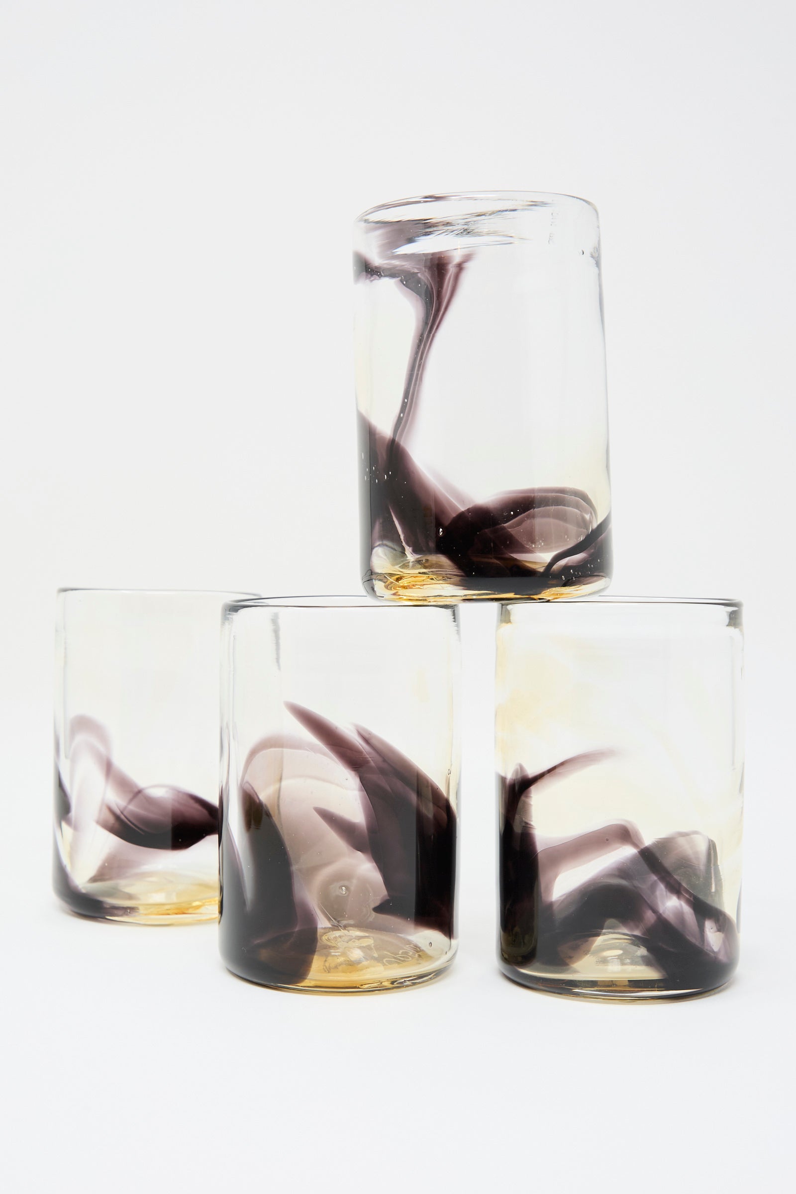 Upstate's Hand Dyed Fumé Glass set features four handmade tumblers with abstract black and brown swirls—three stand upright, while one balances on top, each crafted in NYC.