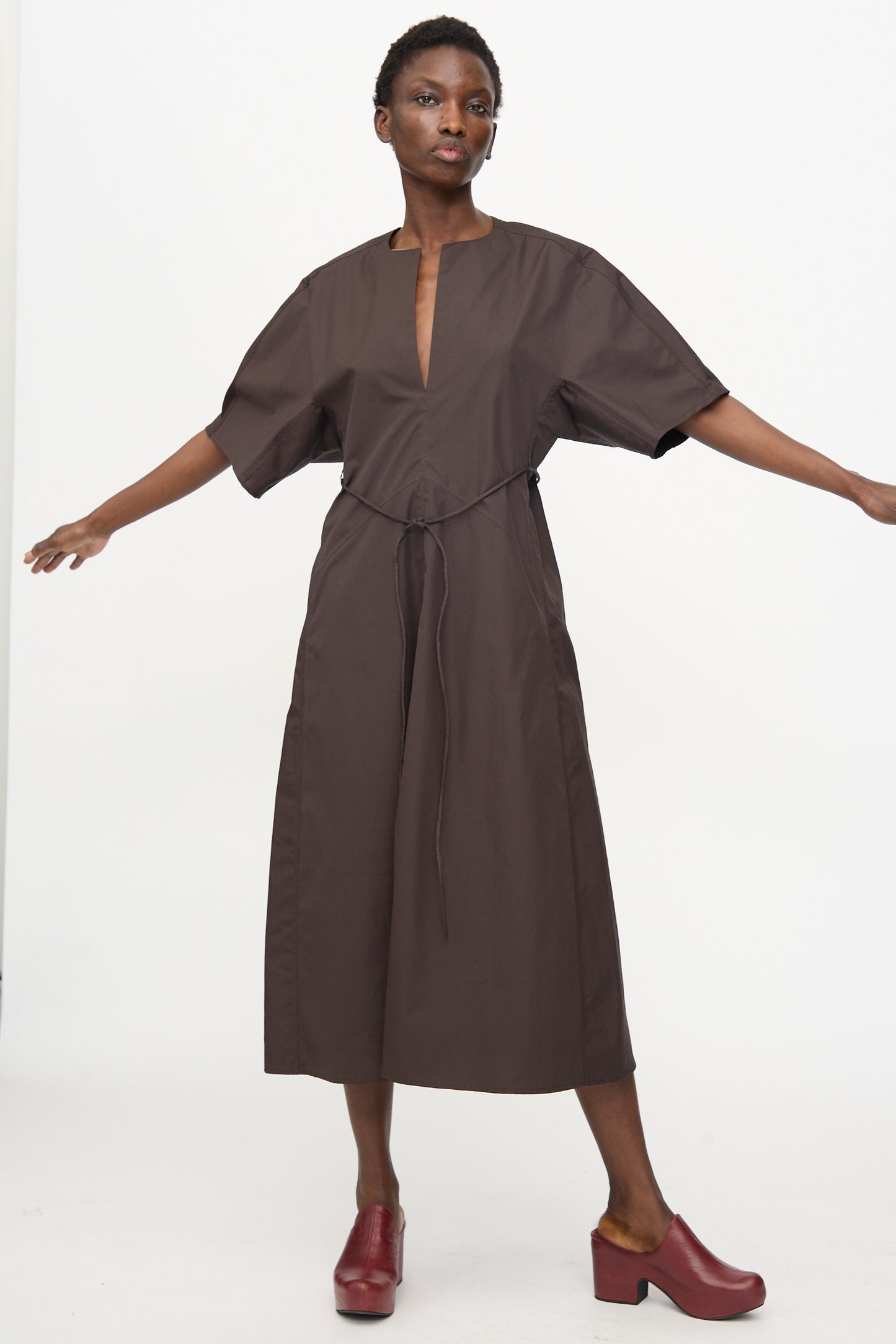 A person stands with arms outstretched, wearing the Veronique Leroy Belted Kimono Dress in Choco—a relaxed fit brown cotton dress with front slit and belt—paired with red heeled clogs against a plain white background.