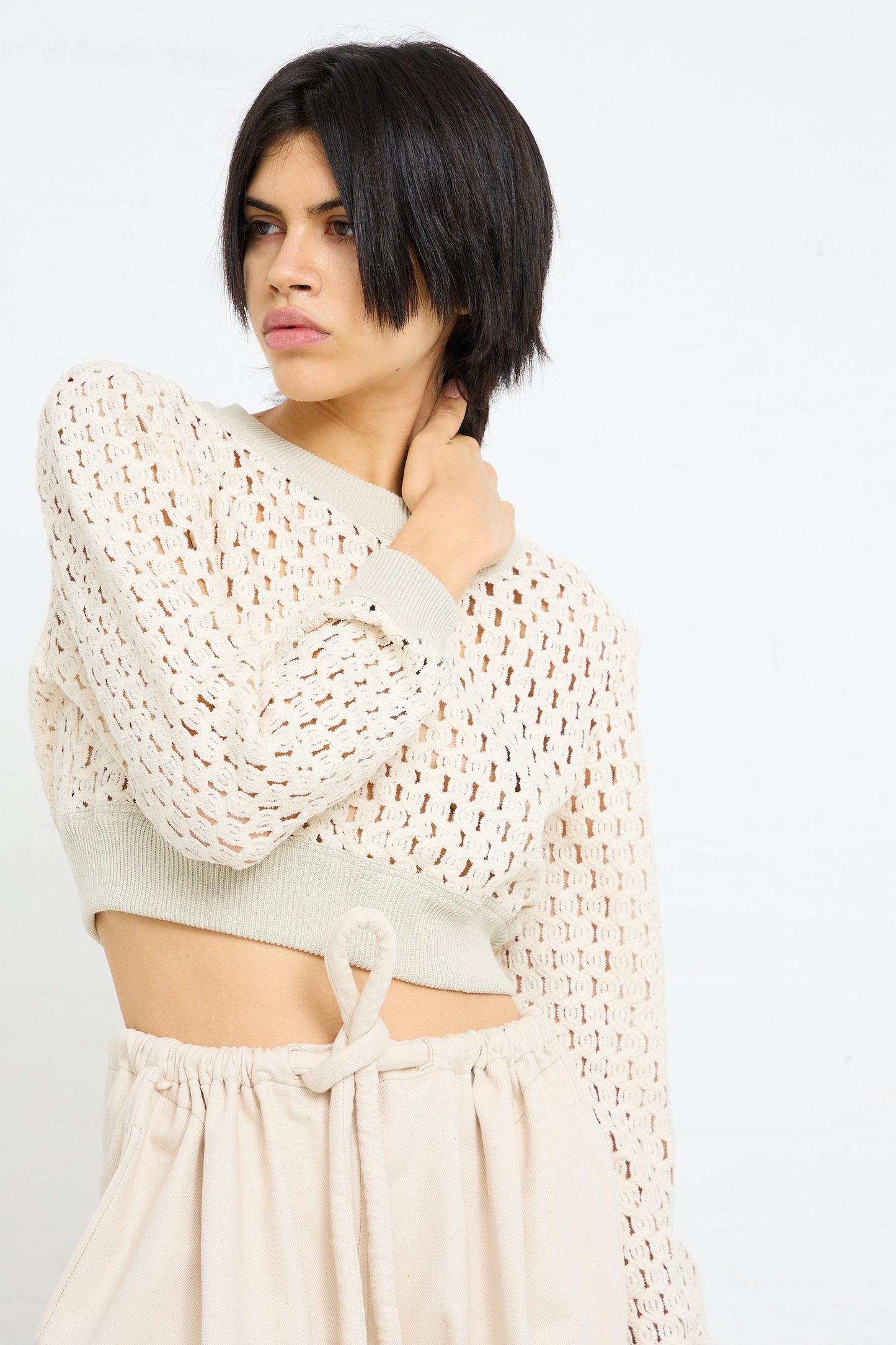 A woman with a short dark bob haircut wearing a Cotton Macro Mesh Cropped Sweater in Ivory by Veronique Leroy and pants, looking to the side with a thoughtful expression.