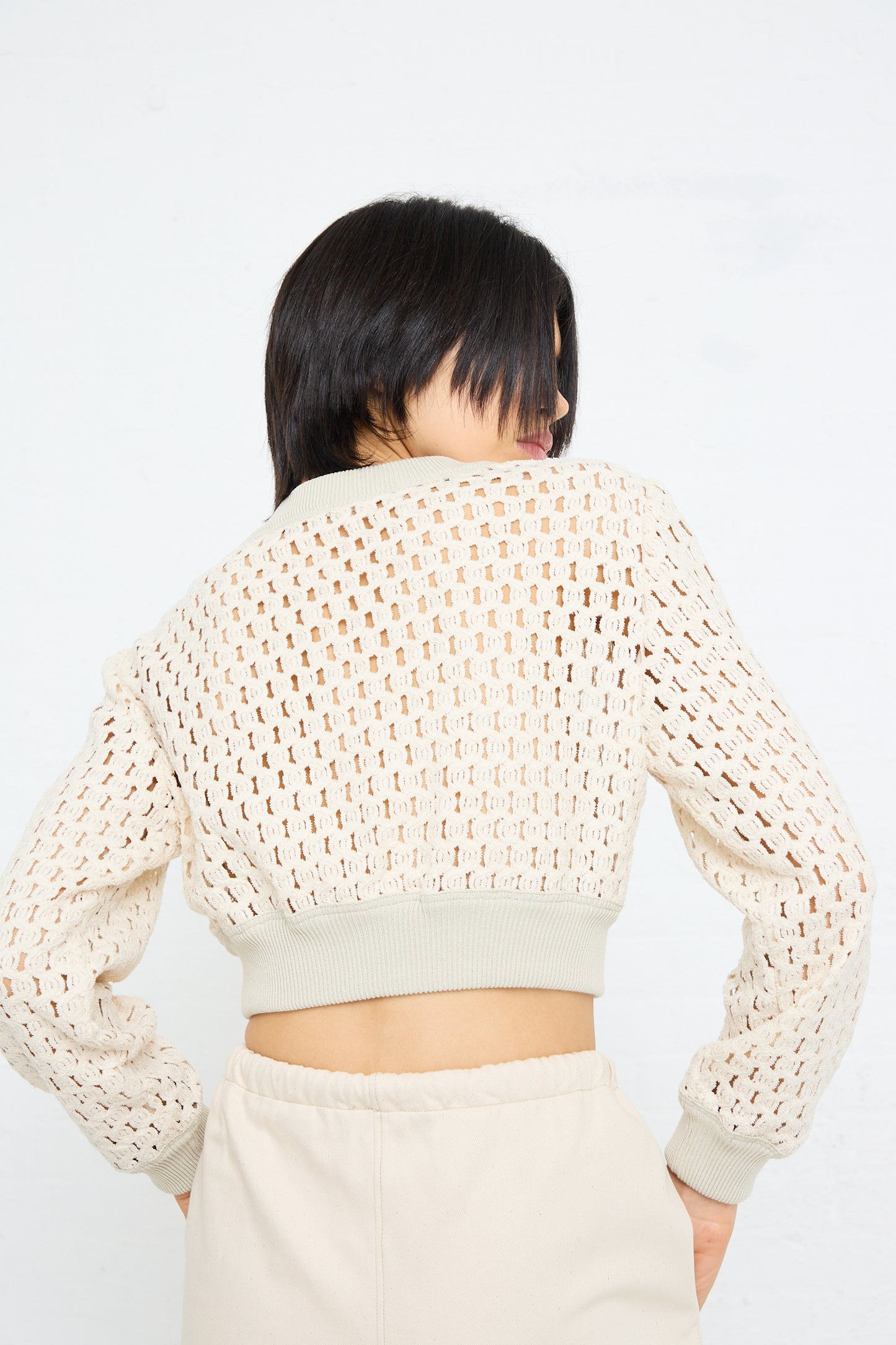 A woman with her back to the camera wearing a Cotton Macro Mesh Cropped Sweater in Ivory by Veronique Leroy with a knit pattern and high-waisted trousers.