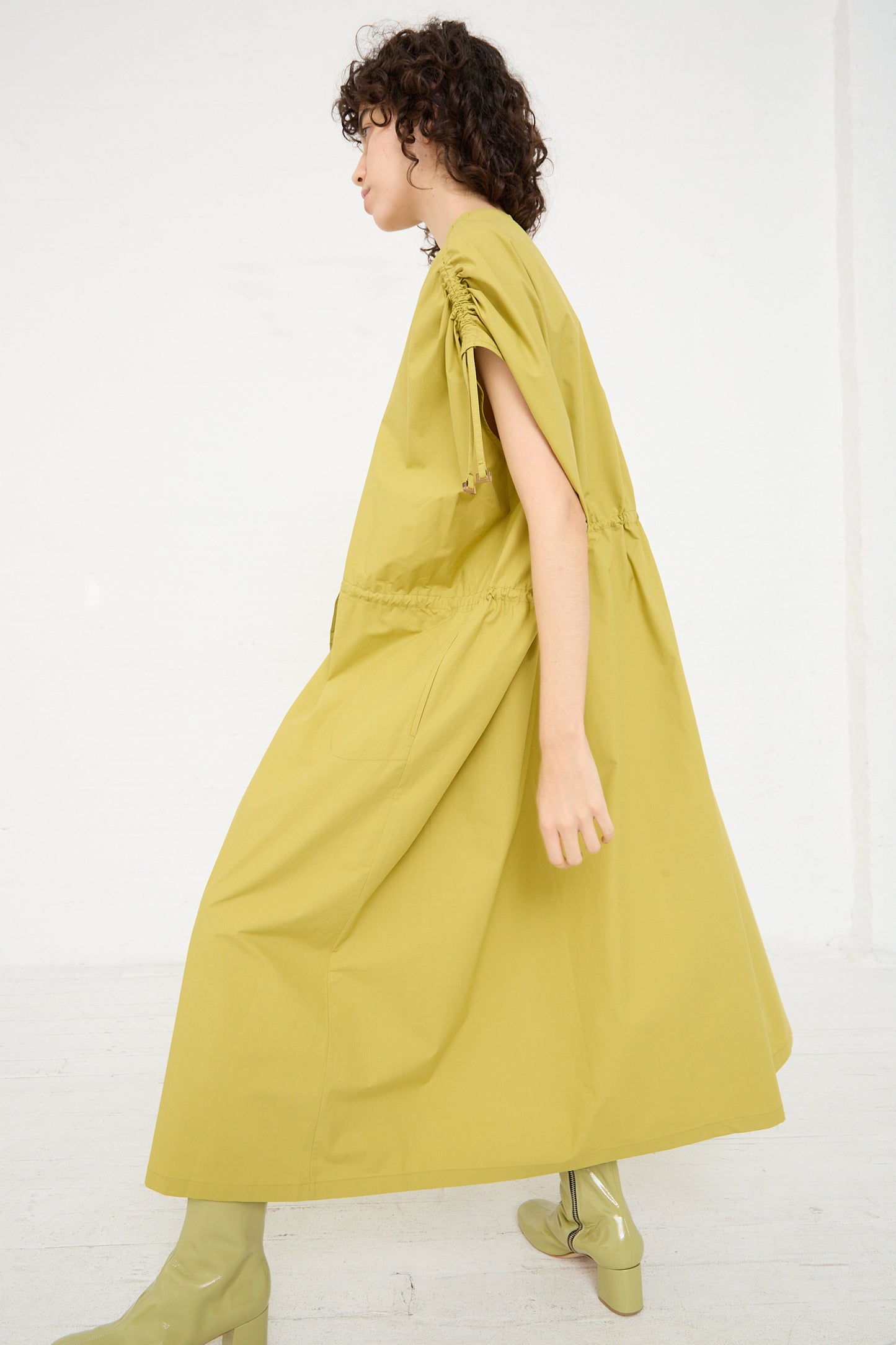A woman wearing a yellow Cotton Poplin Gathered Dress in Matcha by Veronique Leroy and green boots. Side profile.