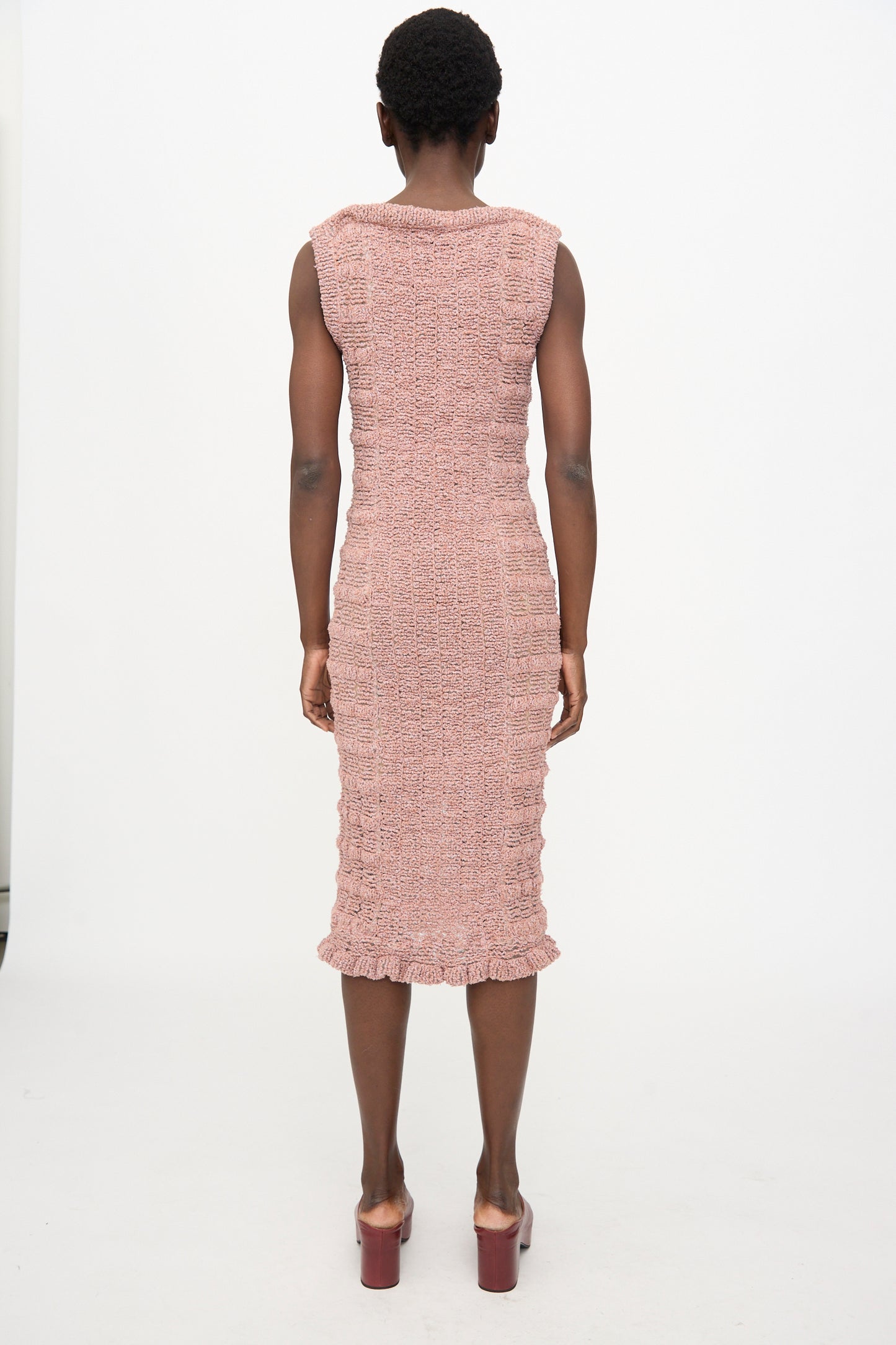 A person stands facing away in the Veronique Leroy Smock Knit Dress in Dusk, a sleeveless cotton-blend midi with a smocked silhouette and ruffled hem, styled with pink heeled sandals against a plain white background.