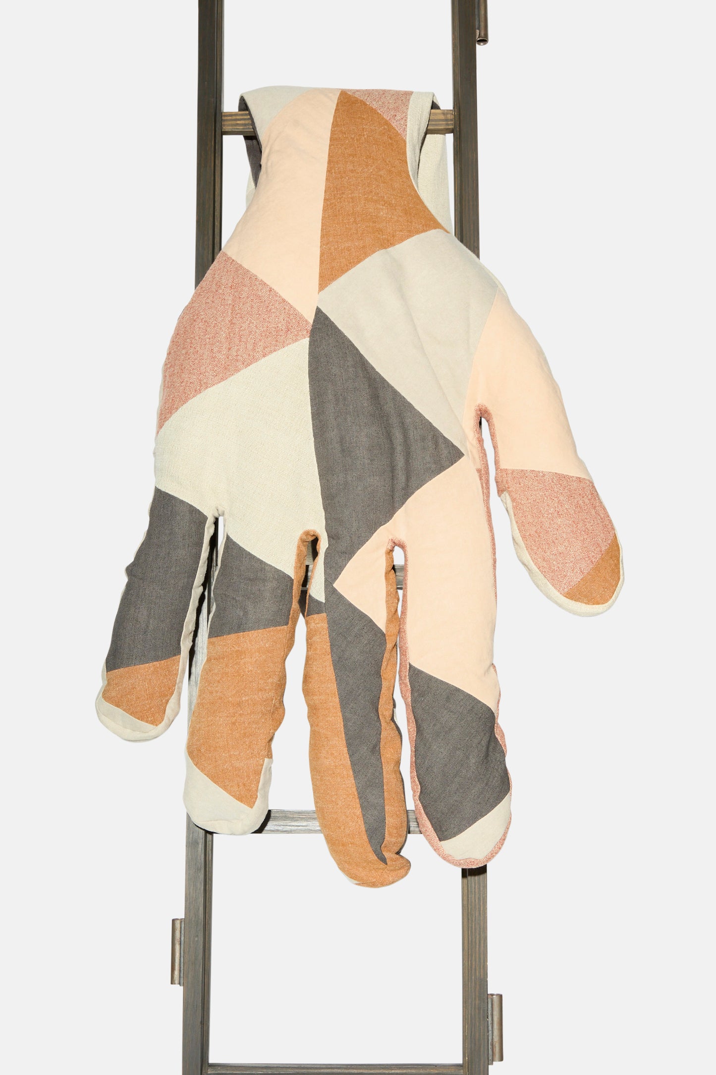 The Sonia Delaunay Tissu Simultané Giant Hand Bolster I by Wiener Times—a large, patchwork hand-shaped pillow inspired by Wiener Werkstätte—drapes over a metal ladder against a plain white background.