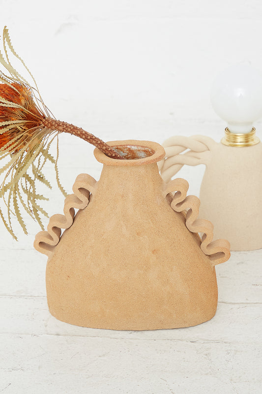 A hand-made ceramic vase with a ruffled design holding dried plants, accompanied by a Clandestine Amphora Soleil vase in Raw Sunny Brown Clay with a bulbous body in the background.