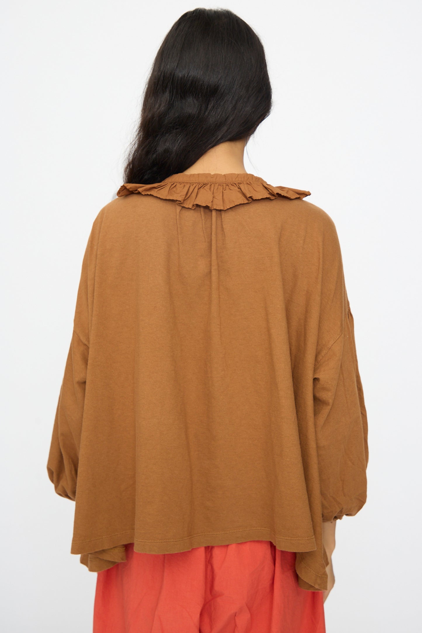 A person facing away wears the nest Robe Cotton Linen Frill Top in Brown with long sleeves and a ruffle collar, paired with a coral skirt.