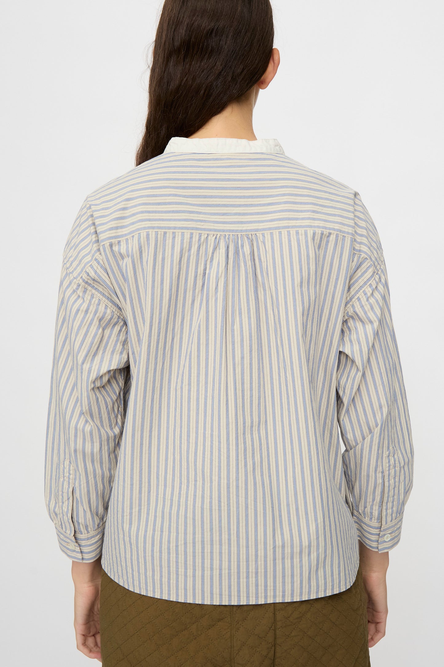 A person with long brown hair wears the nest Robe Cotton Pull-on Shirt in Stripe, a relaxed-fit blue and white vertically striped cotton top, paired with olive quilted pants and shown from the back.