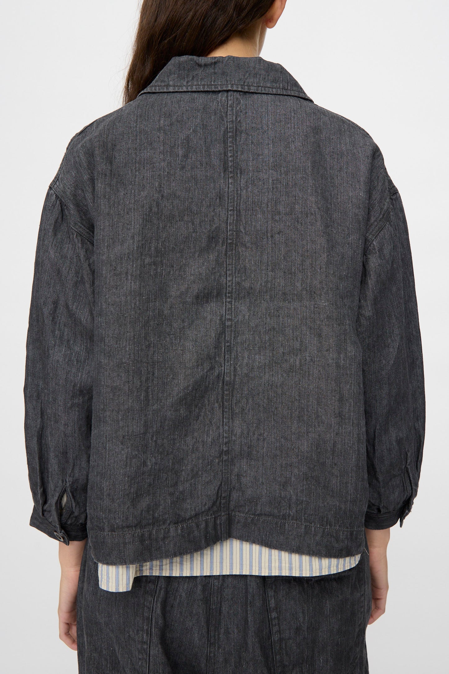 Person wearing the nest Robe Linen Denim Cropped Jacket in Black, featuring a boxy fit and long sleeves over a striped shirt. Shown from the back.