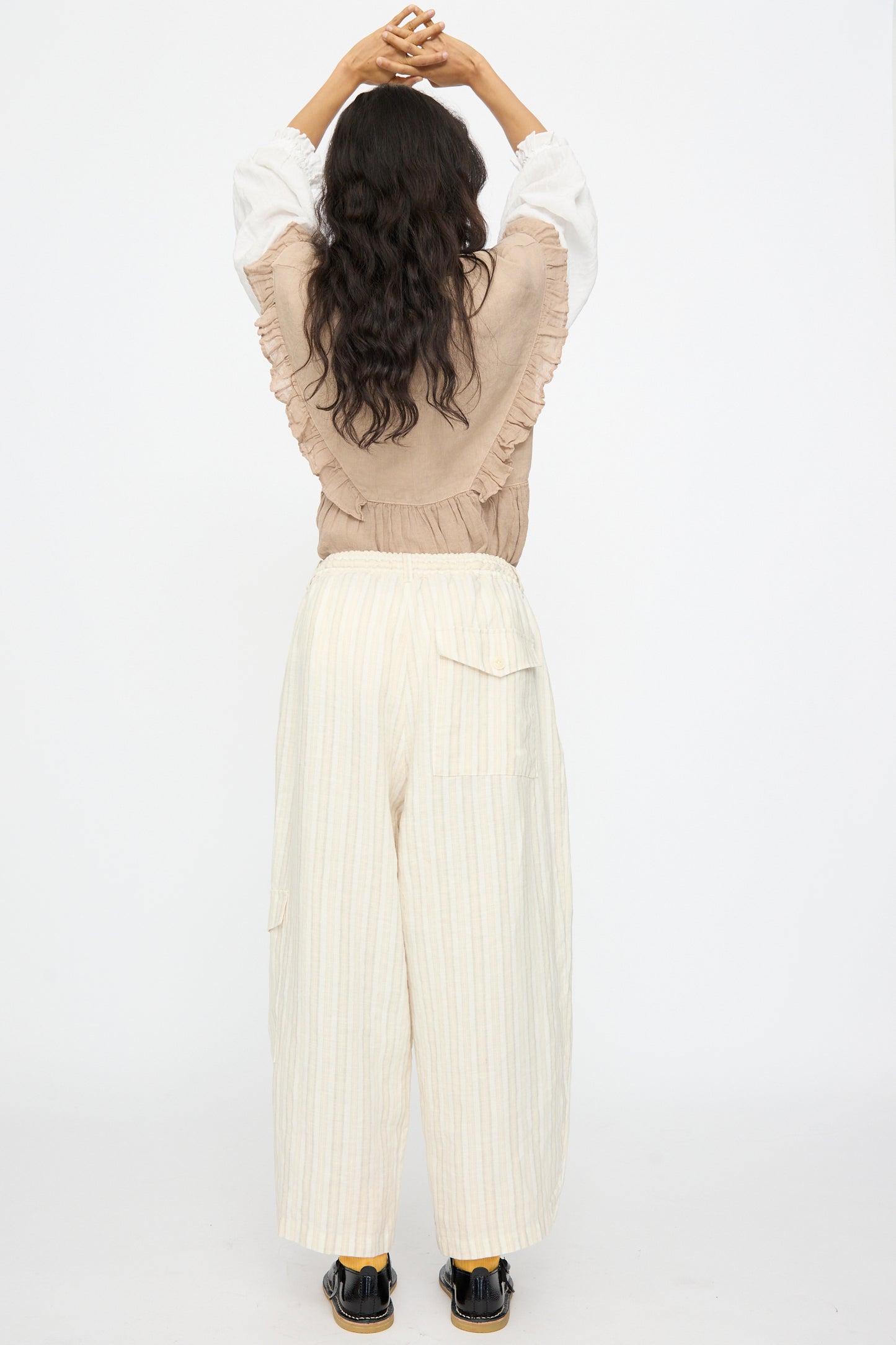 A person stands with arms raised, back to camera, wearing a beige ruffled top and nest Robe’s Linen Easy Pants in Natural Stripe featuring an elastic waistband.