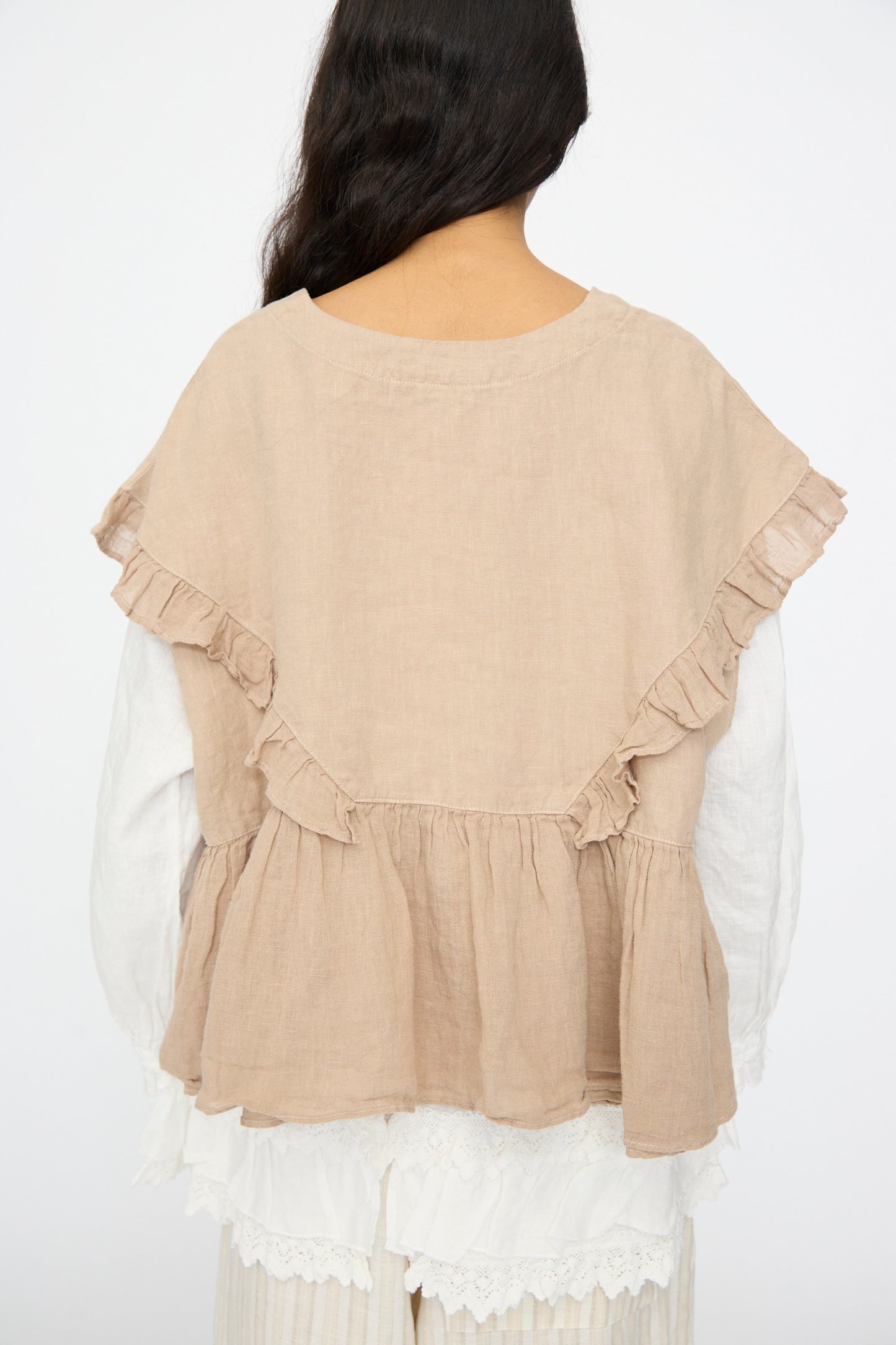 A person with long dark hair is photographed from the back against a plain background, wearing the nest Robe Linen Frill Vest in Beige layered over a white blouse.