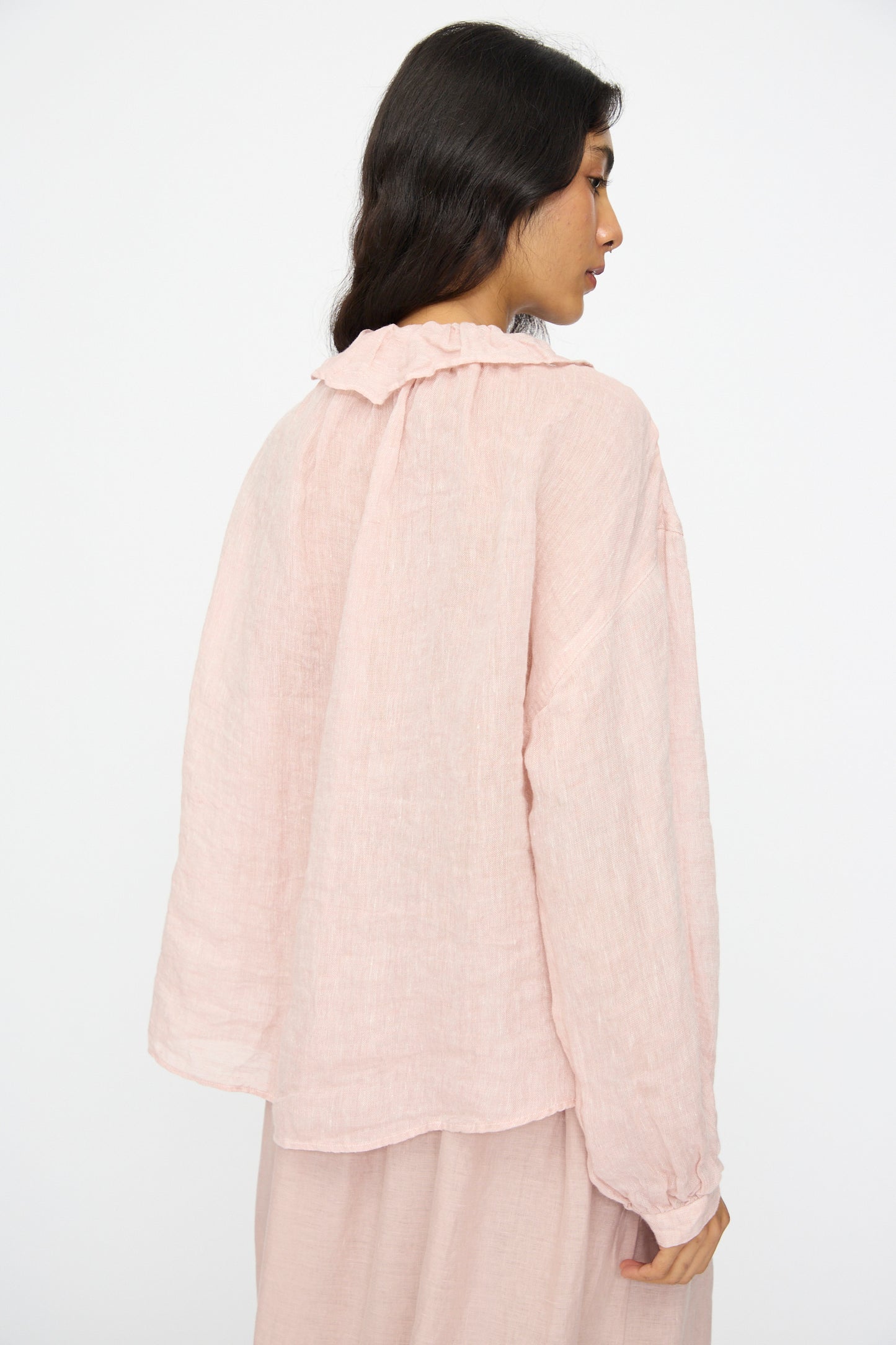A woman with long dark hair models the nest Robe Linen Gauze Frill Blouse in Light Peach, shown loose-fitting with long sleeves, paired with a matching skirt and viewed from the side and back against a white background.