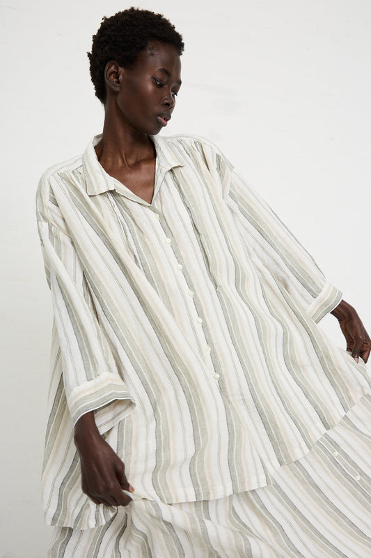 Person wearing a relaxed fit shirt with light and dark vertical lines against a plain background. The loose-fitting, long, striped Linen Long Length Shirt in Stripe by nest Robe features a front button design that adds to its casual elegance.
