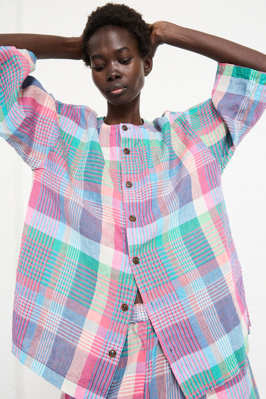 A person with short hair wears a relaxed-fit, multi-colored plaid button-up shirt, the Linen Madras Wide Sleeve Blouse in Check by nest Robe, and poses with their arms raised above their head.