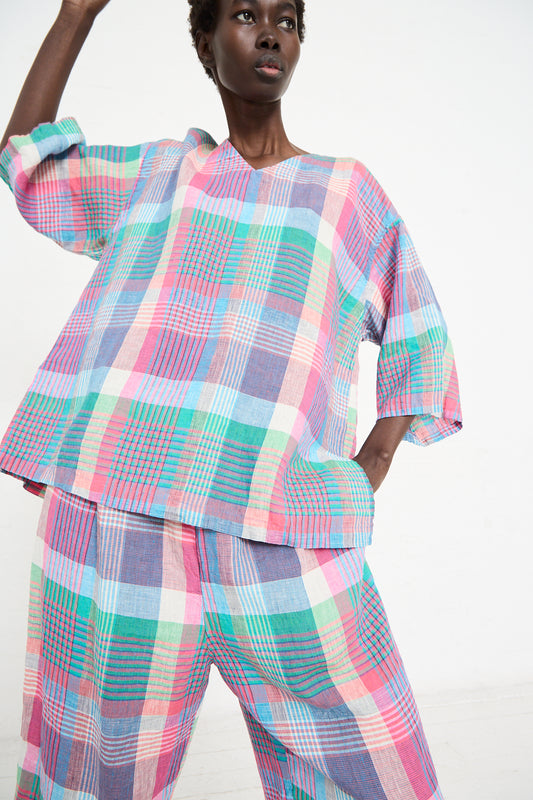 Person wearing a colorful, plaid-patterned outfit, with a relaxed fit Linen Madras Wide Sleeve Blouse in Check by nest Robe, standing with one arm raised and the other hand in their pocket. The background is plain and white.