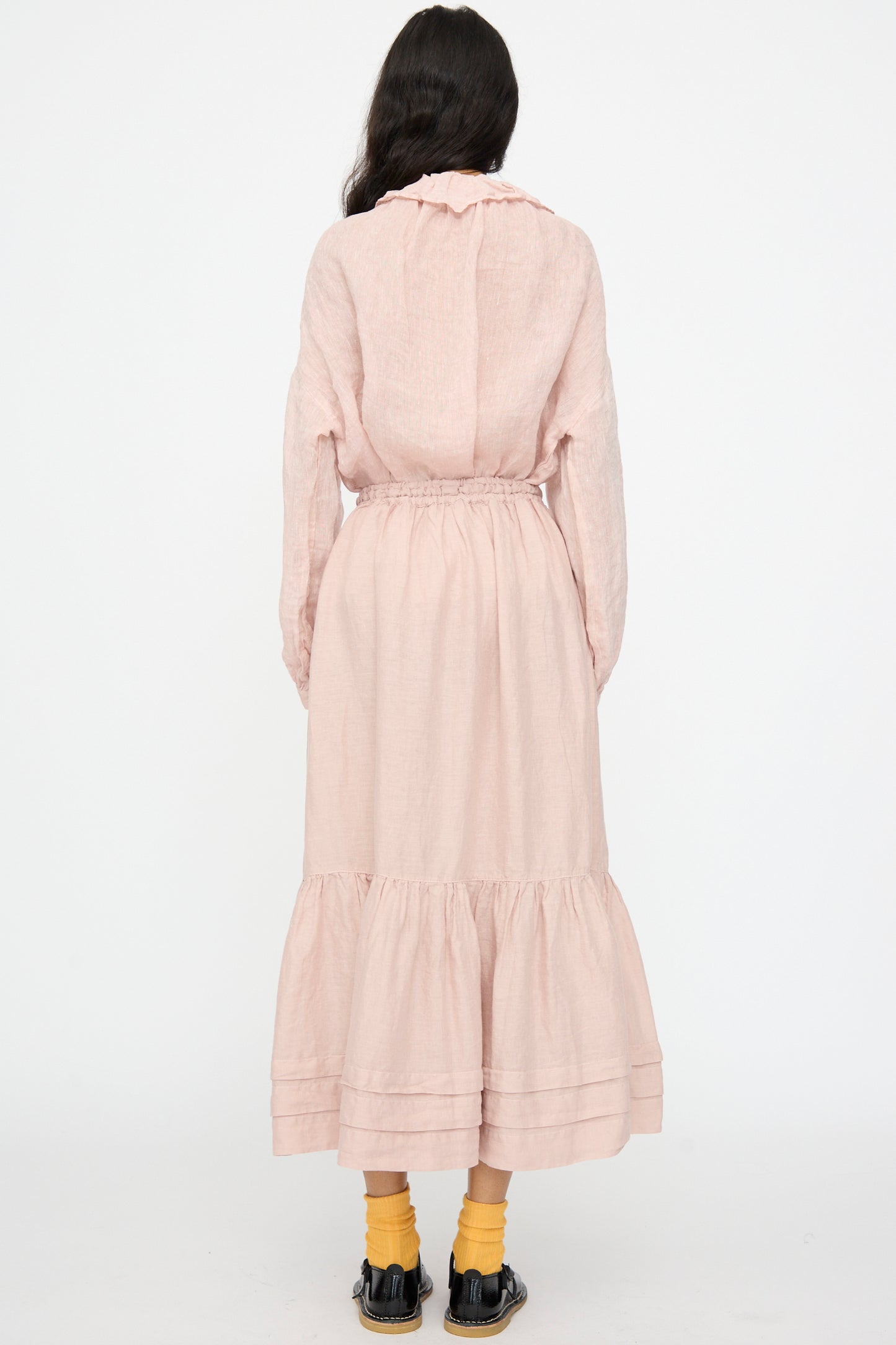 A person stands facing away, wearing the nest Robe Linen Natural Dyed Pleated Gathered Skirt in Pink, a light pink long-sleeve dress with a ruffled hem, yellow socks, and black shoes against a plain white background.
