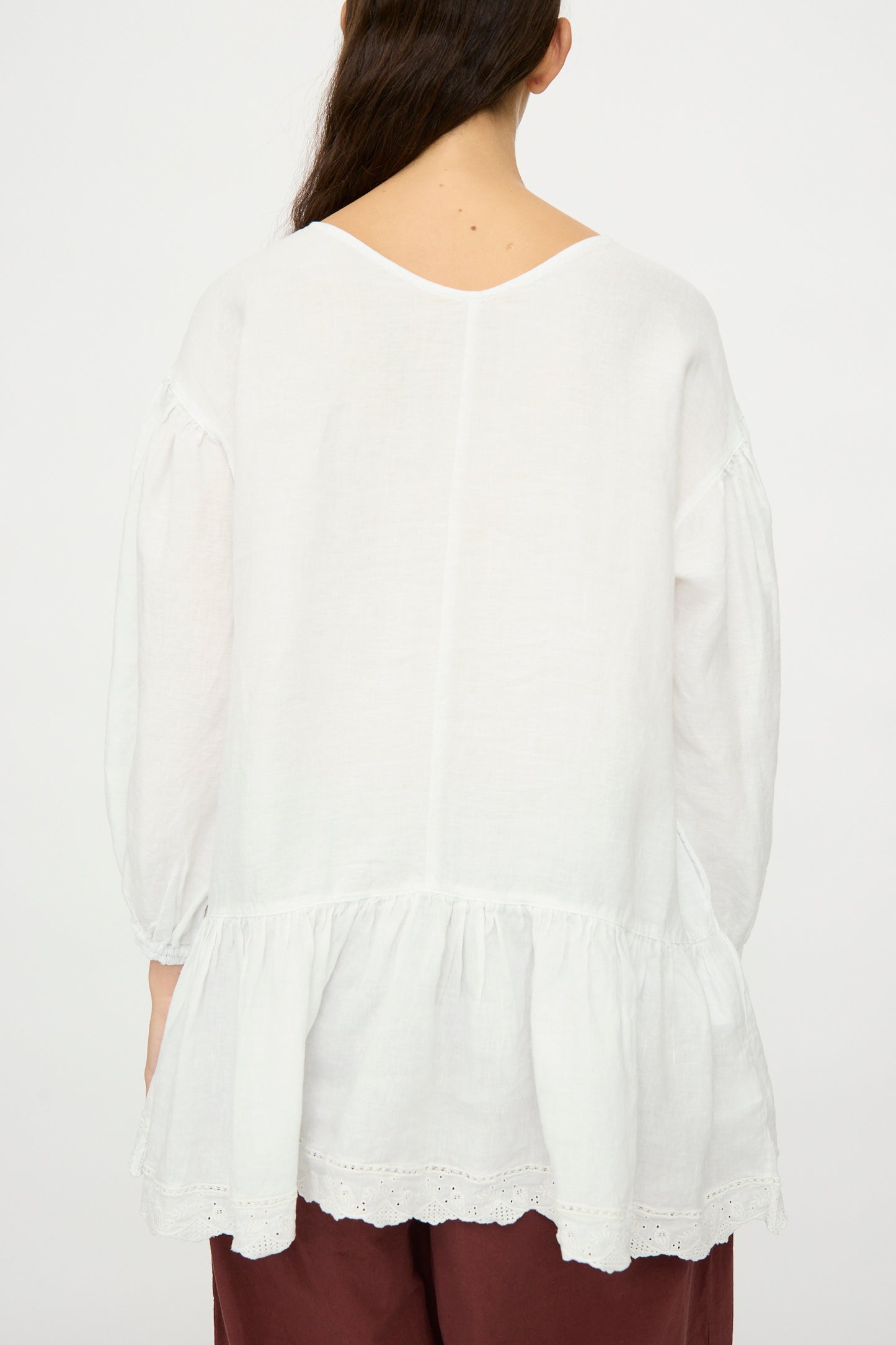 A person with long dark hair, seen from behind, wears the nest Robe Linen Scallop Lace Two-Way Tunic—loose-fitting with bishop sleeves and gathered hem—paired with maroon pants against a plain background.