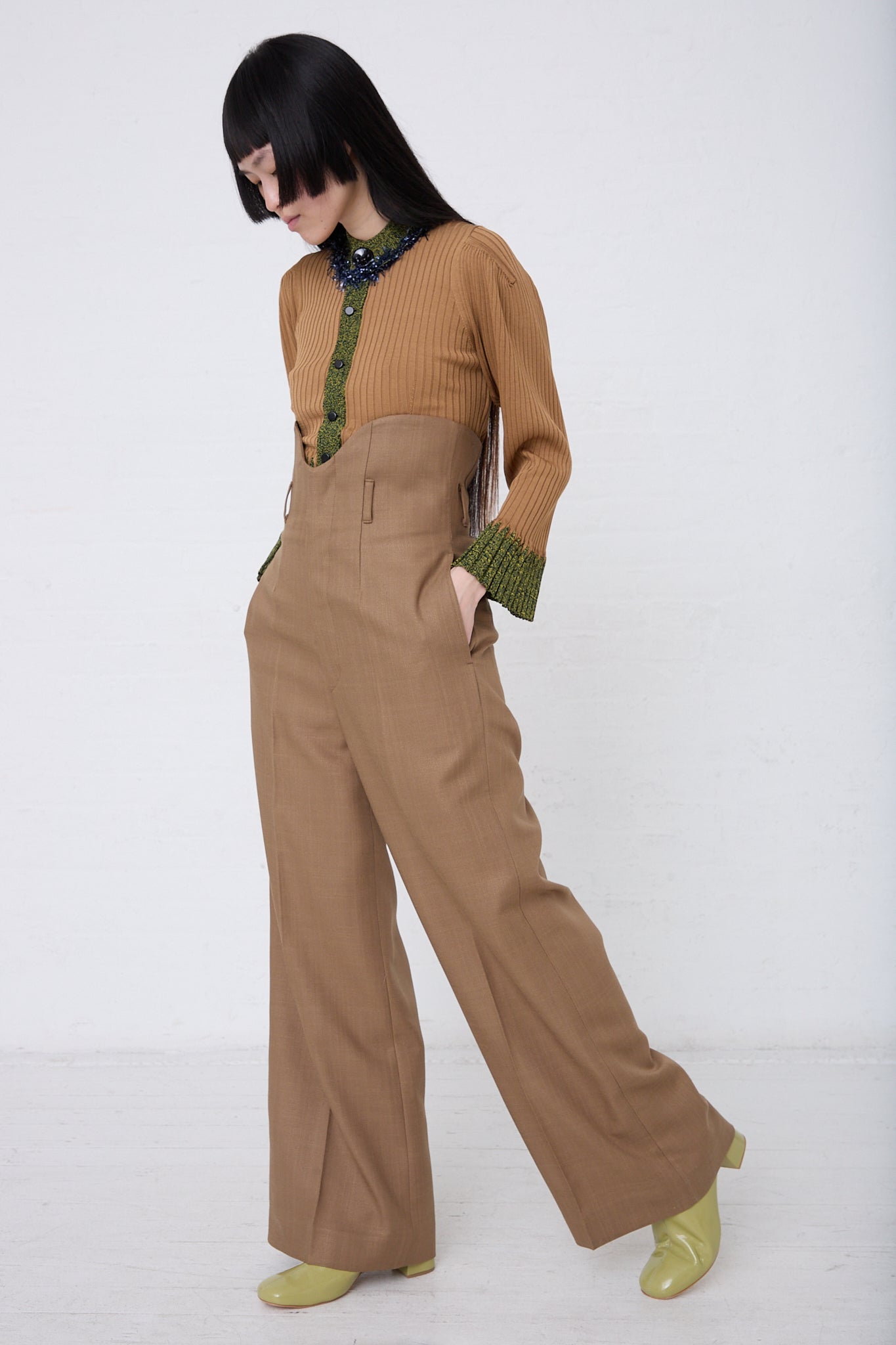A woman in a stylish TOGA ARCHIVES brown pantsuit with a ribbed top and high waist trousers, paired with green gloves and yellow shoes, standing against a plain white background.