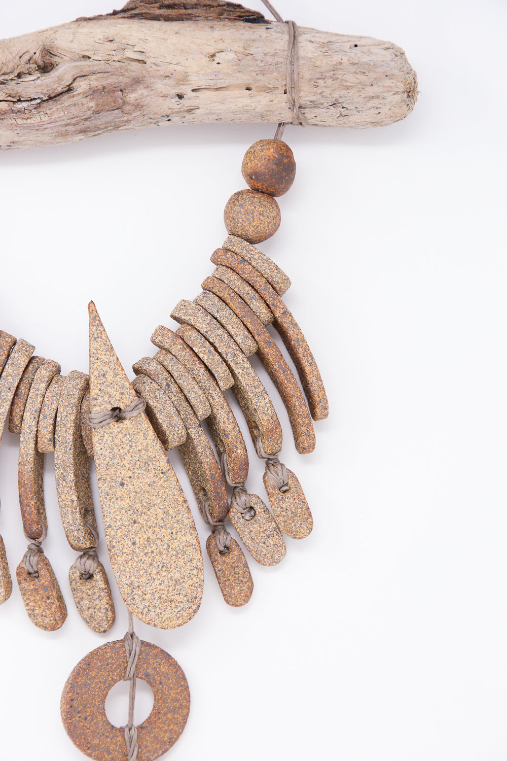 The Heather Levine Teardrop and Circle Wall Hanging in Mixed Brown, featuring large, textured, stone-like beads in various shapes, is beautifully displayed hanging from a piece of driftwood against a white background.