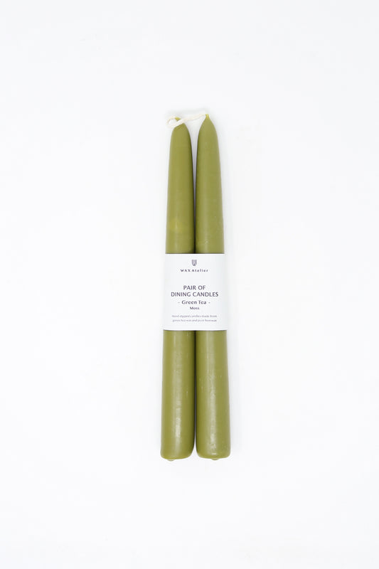 A pair of green Wax Atelier Beeswax Dining Candles in Moss with a label against a white background.