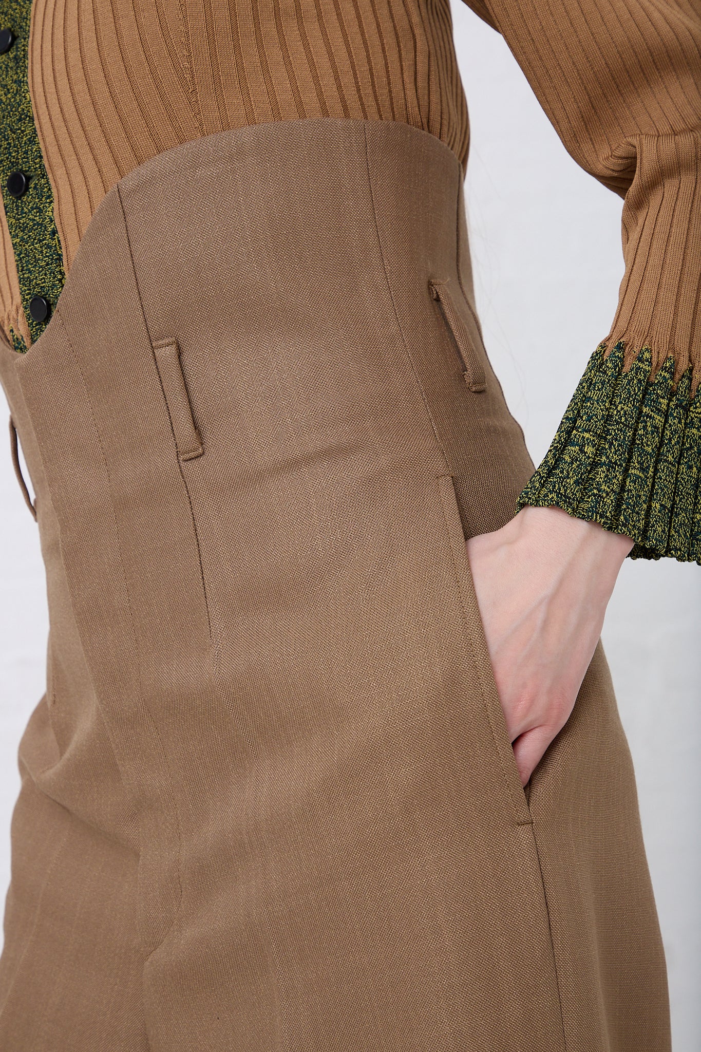 Close-up of a woman's side, showing her hand in the pocket of her TOGA ARCHIVES high waist trouser in Brown with visible stitching and a green blouse with intricate sleeve details.